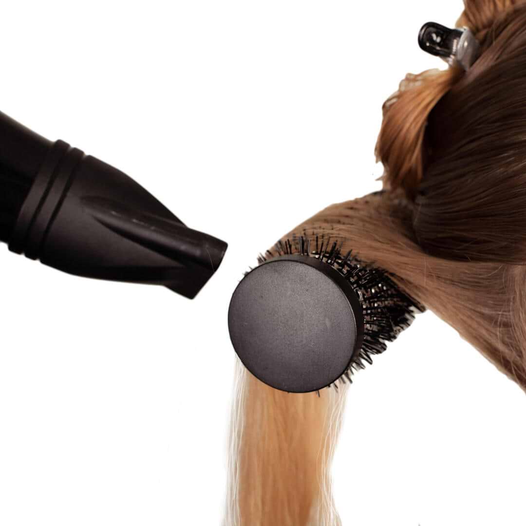 Hair extensions being styled with a round brush and blow dryer, focusing on a section of light brown hair.