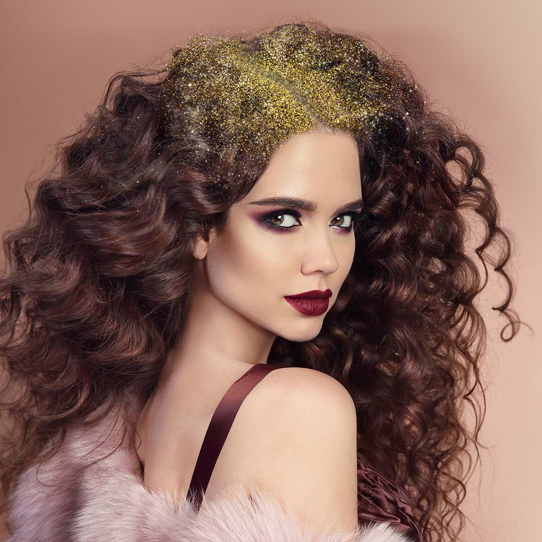 Woman with voluminous curly hair extensions adorned with gold glitter, wearing dark makeup and a fur-like garment, poses against a soft brown background.