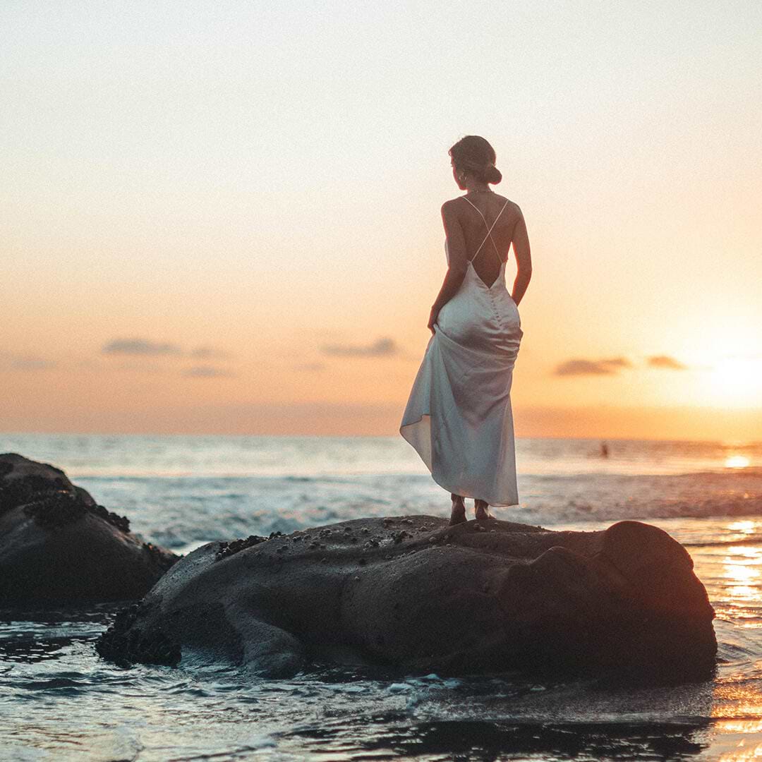 Woman in a white dress stands on a rock by the ocean, gazing at a serene sunset with a colorful sky.