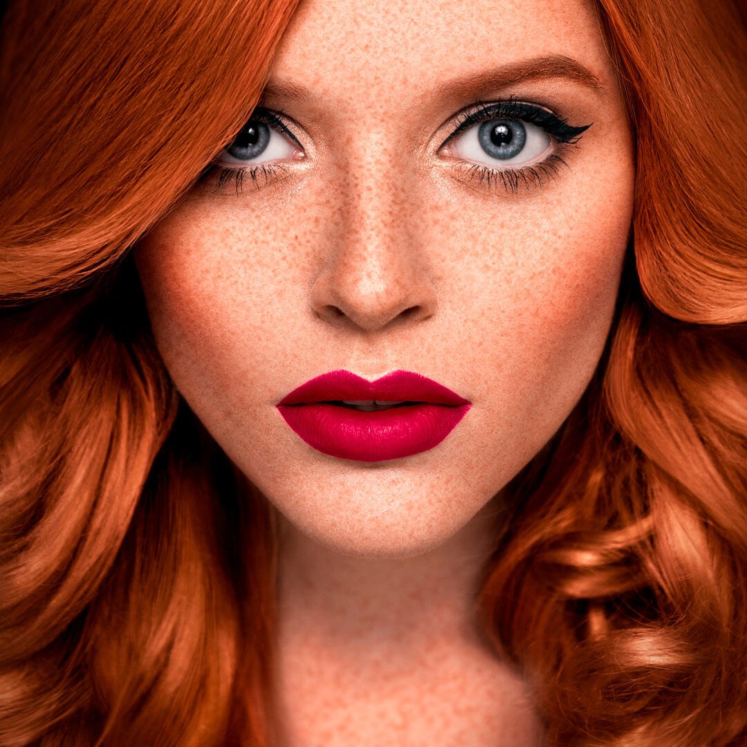 Close-up of a person with vibrant red hair extensions, striking blue eyes, bold red lipstick, and freckles, gazing directly at the camera.