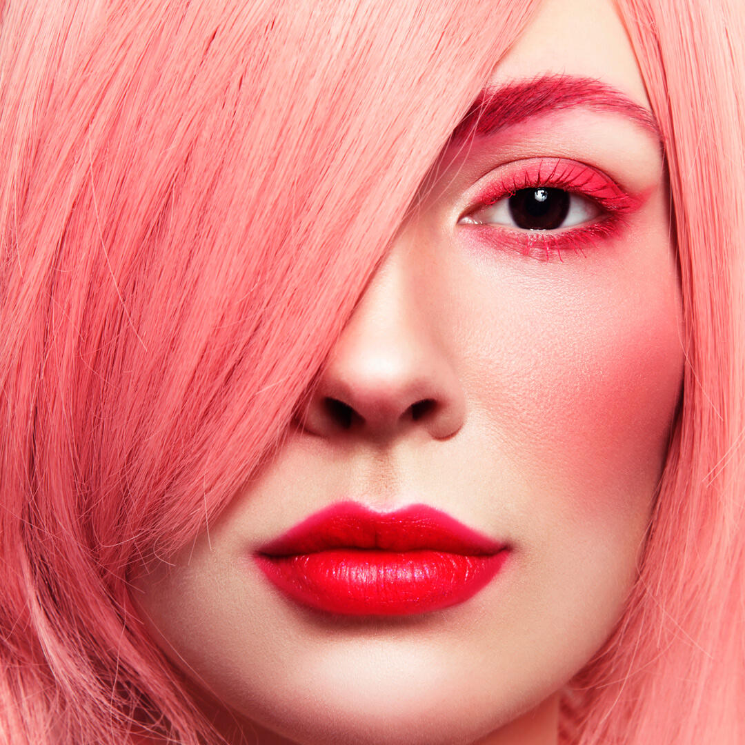 Close-up of a person with pink hair extensions and makeup, featuring vibrant pink eyeshadow, blush, and red lipstick, partially covering one eye.