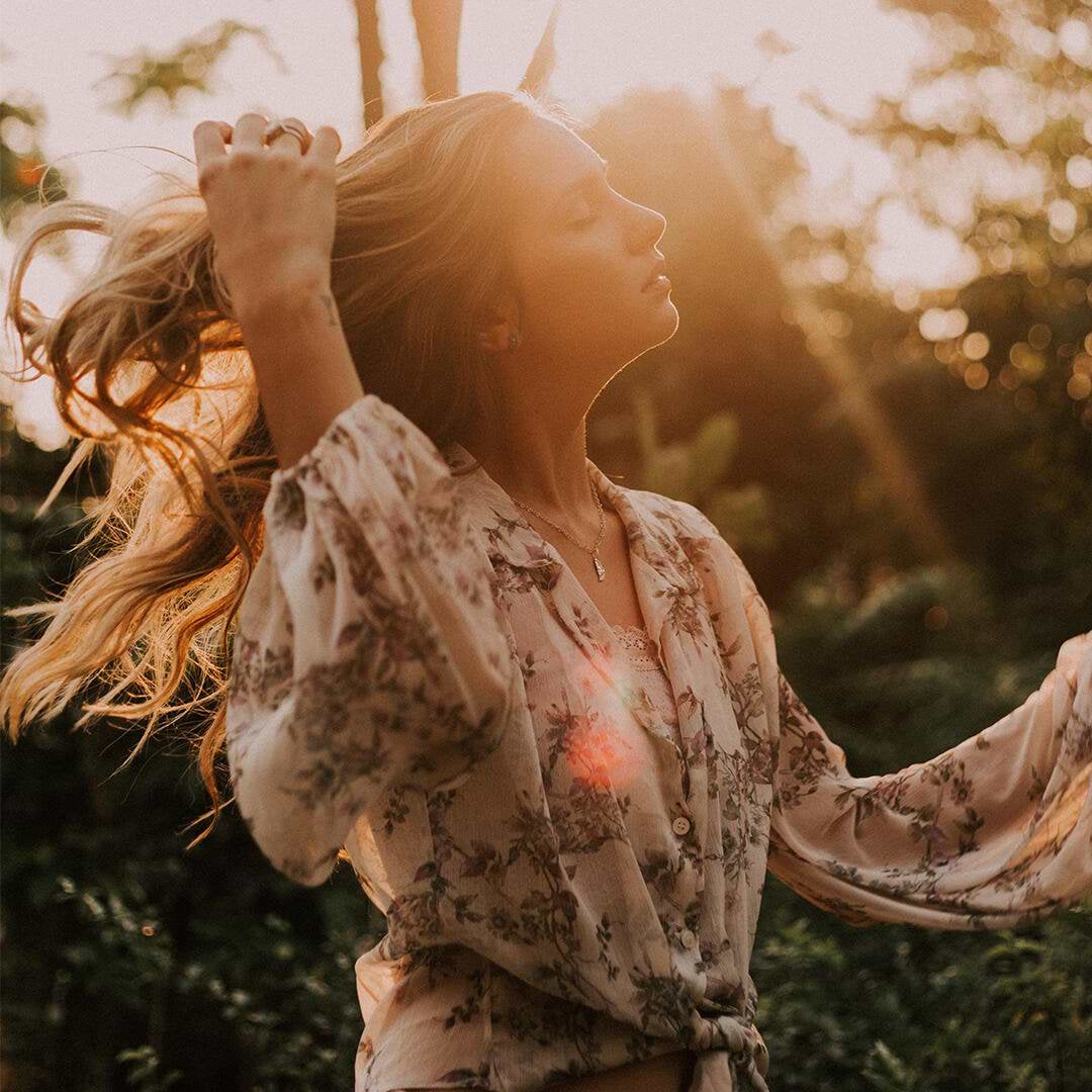 Woman in a floral blouse stands outdoors, eyes closed, with sunlight filtering through her hair extensions, surrounded by greenery.