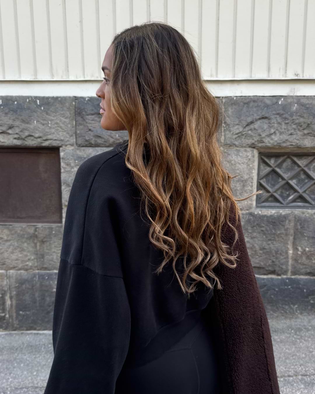Woman with long, wavy brown hair stands outdoors, wearing a black outfit. She is facing away, with a stone wall in the background.