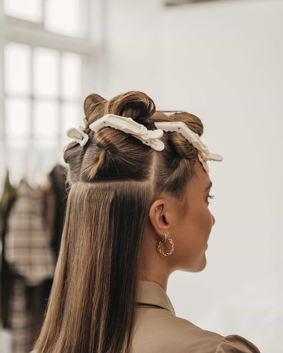 Woman with hair sectioned and clipped, preparing for hair extensions and styling. She wears hoop earrings and a beige top, standing in a bright room.