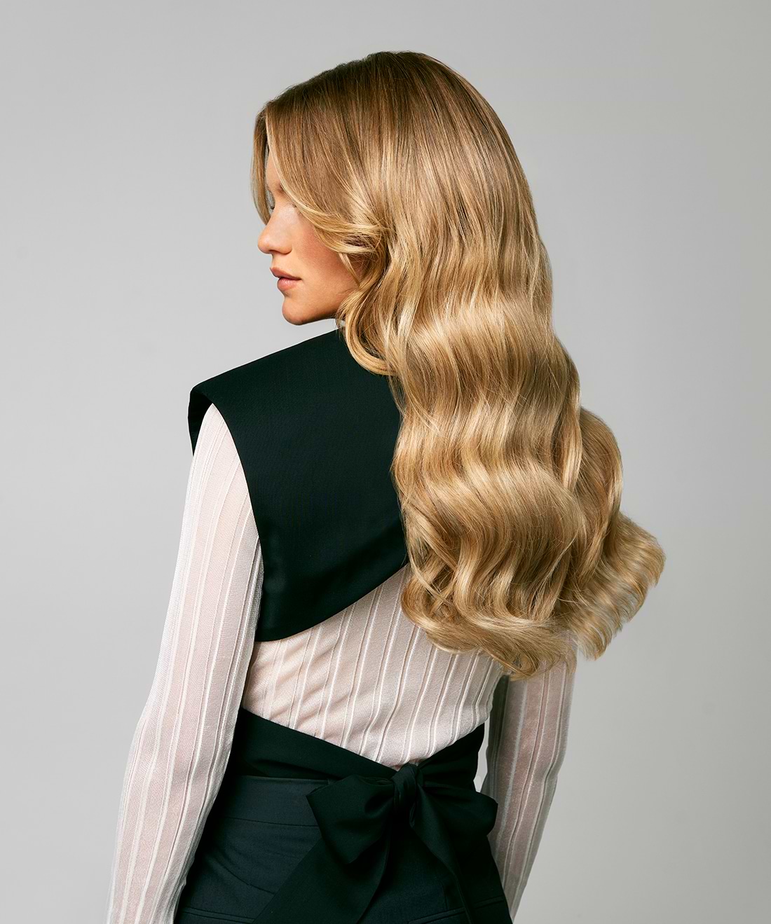 Woman with long, wavy blonde hair extensions wearing a white blouse and black vest, viewed from the side against a plain background.