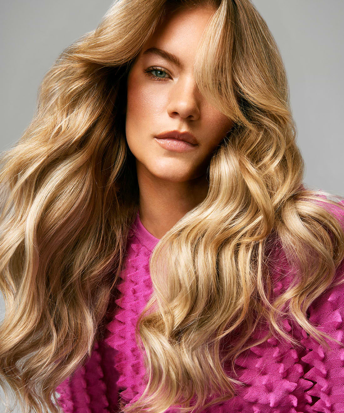 Woman with long, wavy blonde hair extensions wearing a textured pink top, looking confidently at the camera against a neutral background.
