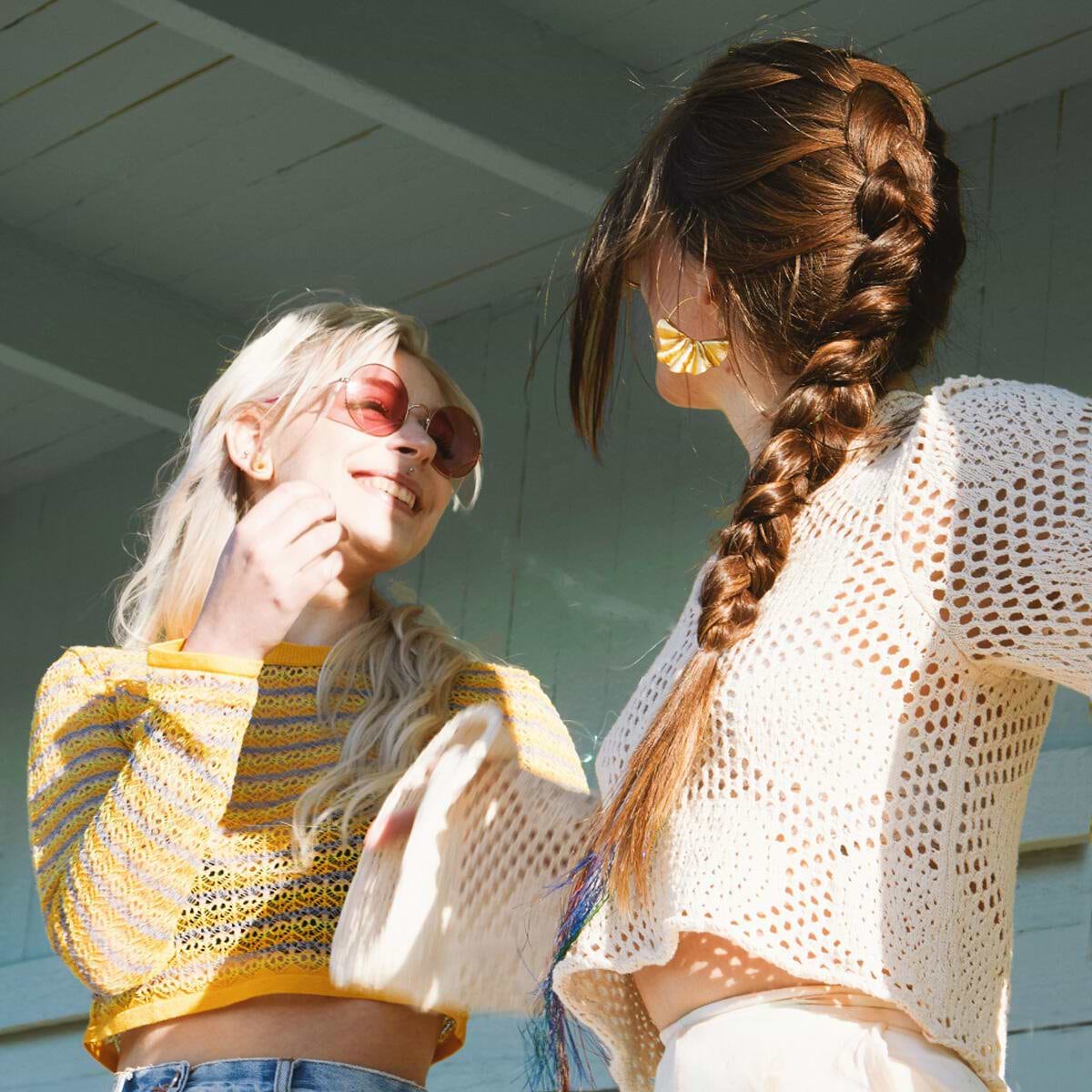 Two women in crocheted tops laughing outdoors. One wears pink sunglasses, the other has a long hair extensions in a braid. Bright sunlight enhances the scene.
