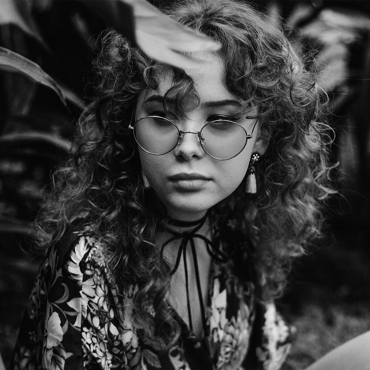 Woman with curly hair extensions and round glasses, wearing a floral outfit, sits outdoors. Black and white image with a contemplative expression.