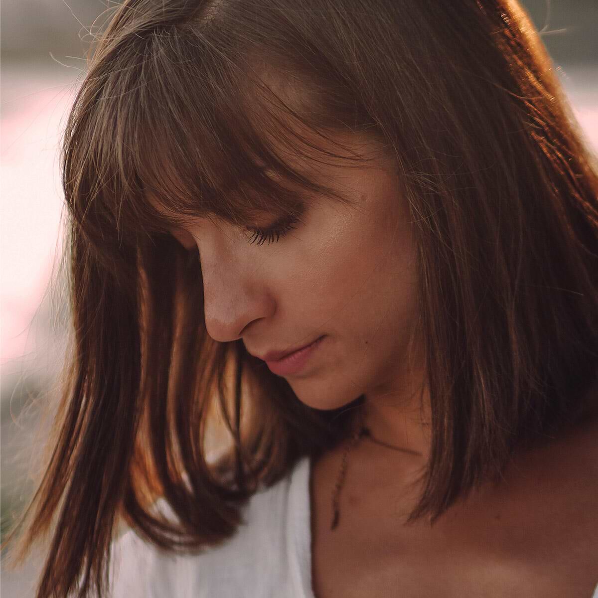 Woman with shoulder-length brown hair extensions and bangs looking down, wearing a white top, softly lit by warm light.