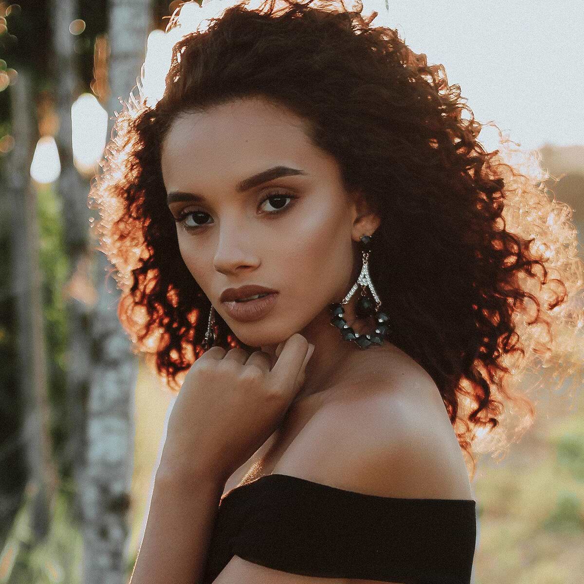 Woman with curly hair extensions and statement earrings poses outdoors, wearing an off-the-shoulder black top, with sunlight illuminating her hair.