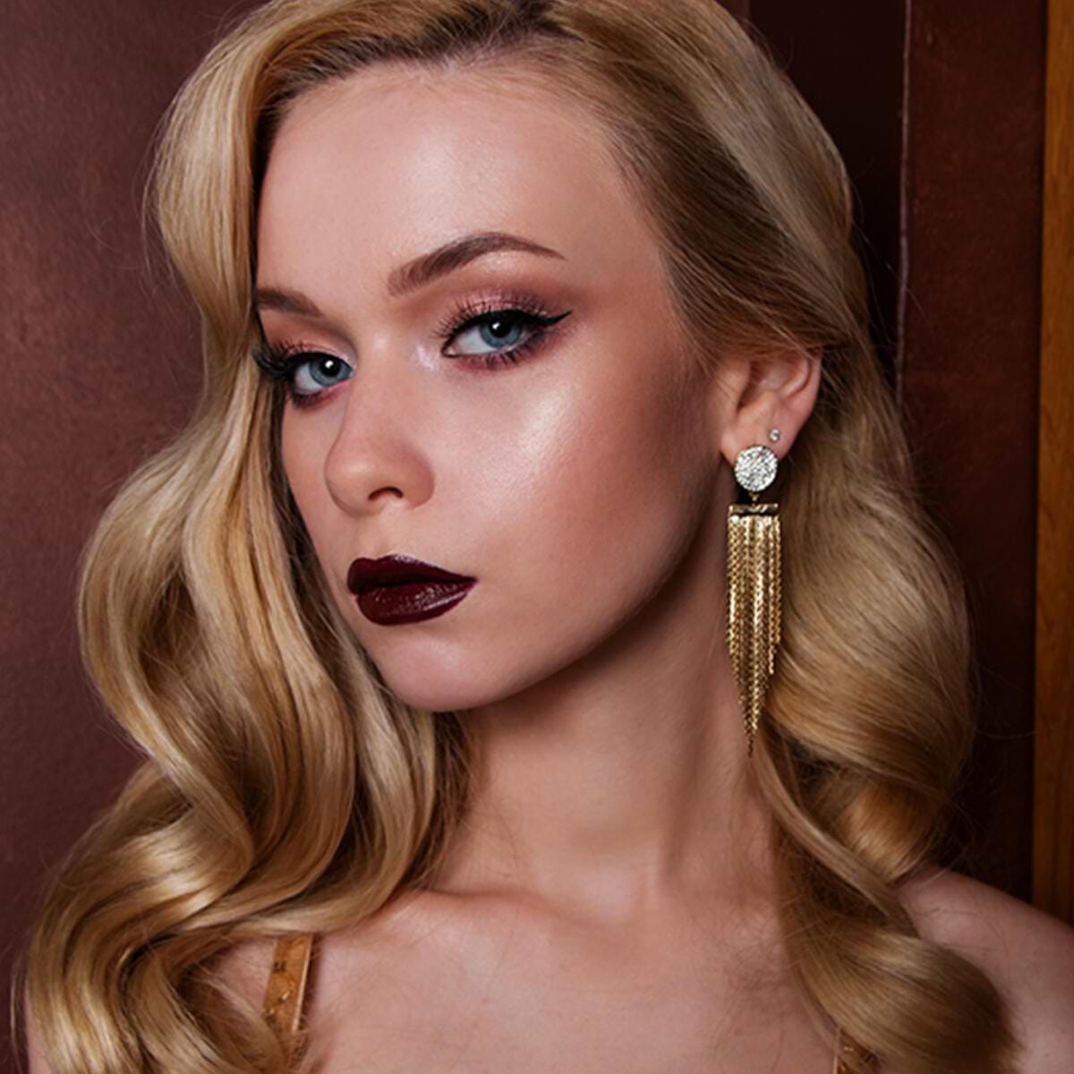 Woman with long blonde hair extensions, dark lipstick, and gold chandelier earrings, posing against a brown background.