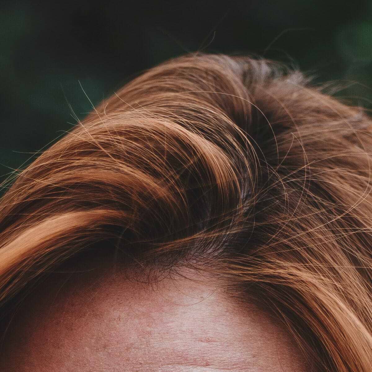 Close-up of the top of a person's head with wavy, light brown hair against a blurred dark background.