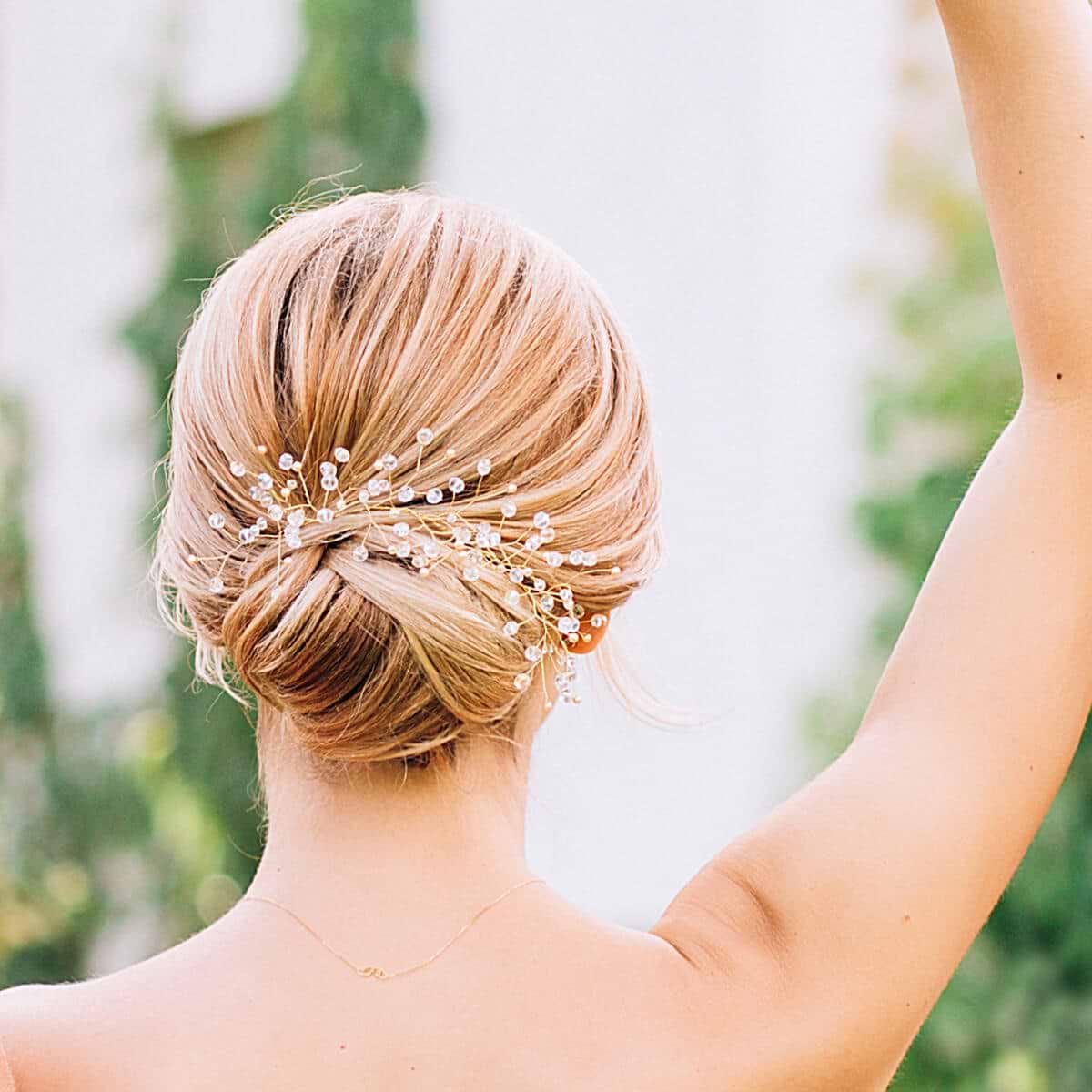 Blonde hair styled in an elegant updo with hair extensions and with delicate pearl and crystal hairpins, shown from the back against a blurred green background.