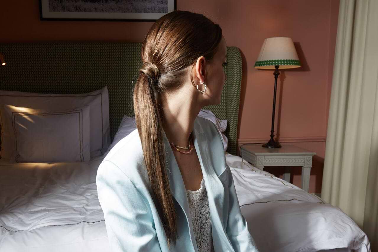A woman with hair extensions in a sleek ponytail sits on a bed, wearing a light blue blazer and earrings, looking to the side in a softly lit room.