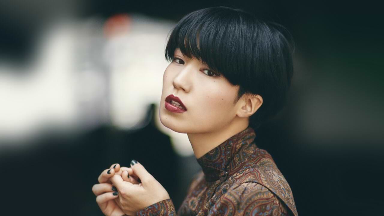Person with short black hair extensions and patterned top, gazing intently at the camera against a blurred background.