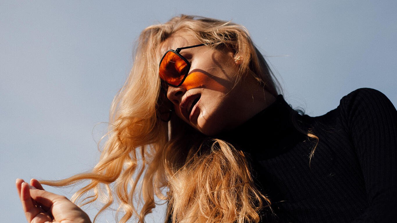 Woman with long blonde hair extensions wearing orange sunglasses and a black turtleneck, looking upwards against a clear blue sky.