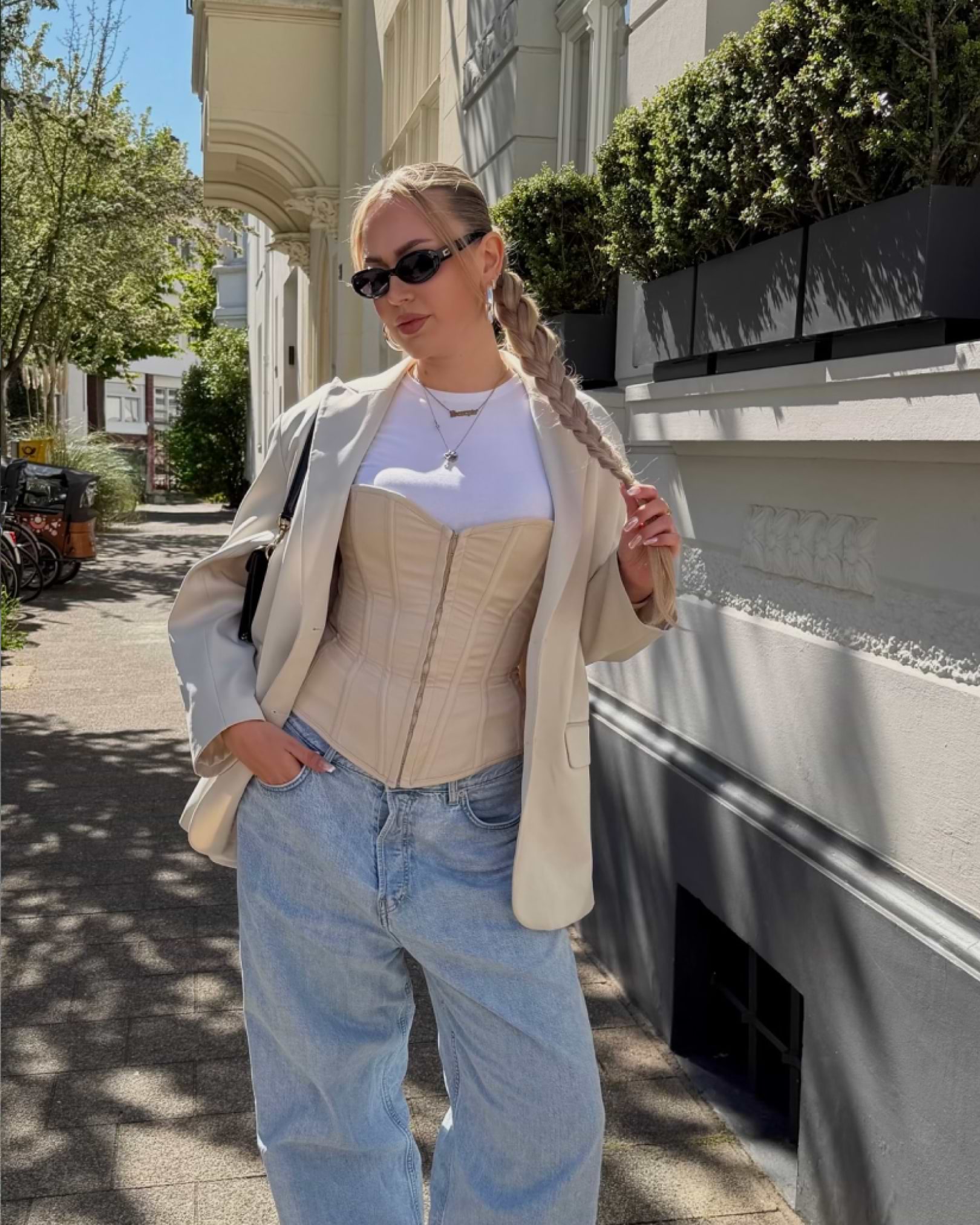 Woman in sunglasses, beige blazer, and corset top stands on a sunlit street. She's wearing wide-leg jeans and has braided hair.