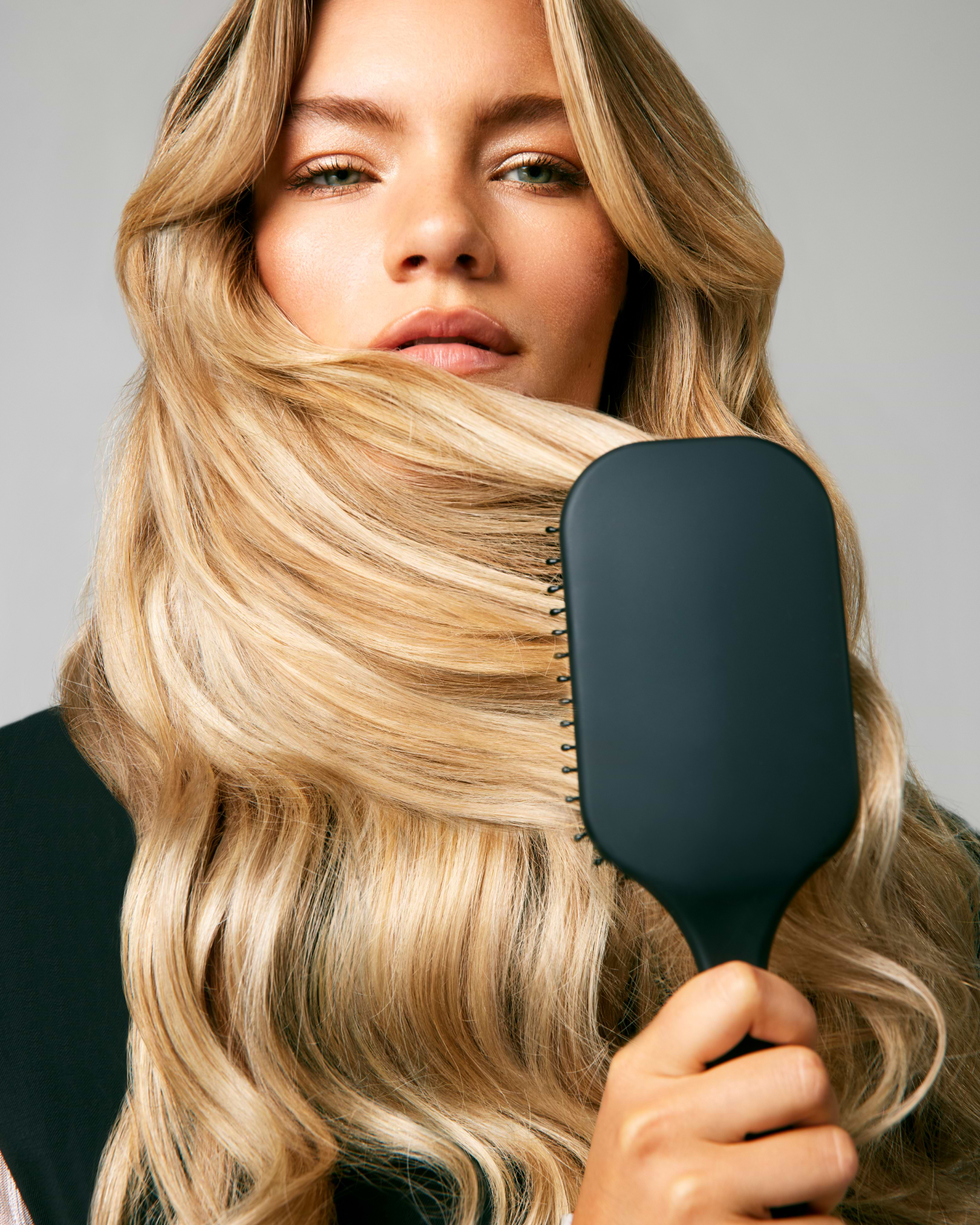 A woman with long, wavy blonde hair holds a black paddle brush in front of her face, highlighting her smooth, shiny hair.