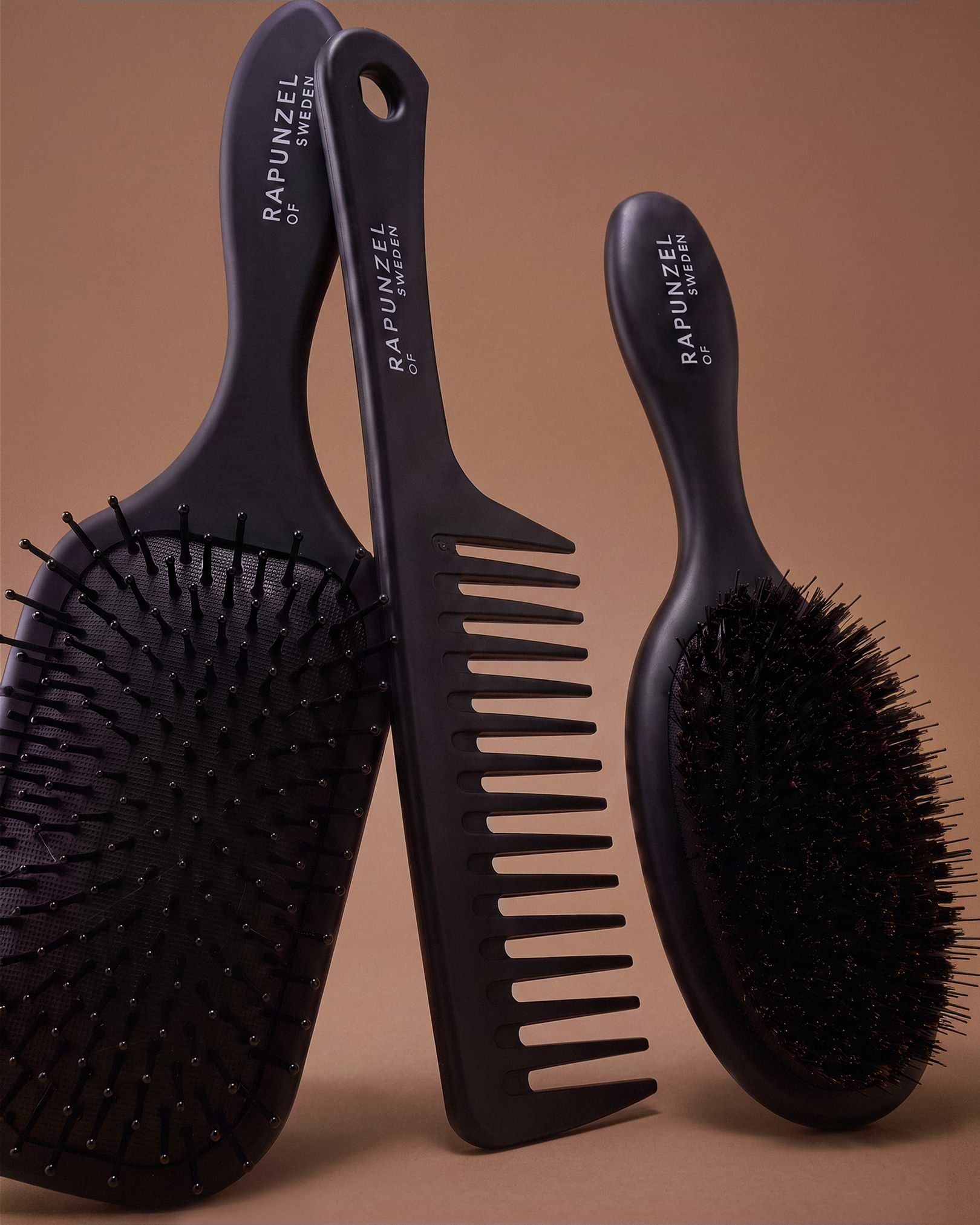 Three black hair tools, including a paddle brush, wide-tooth comb, and round brush, stand against a brown background.