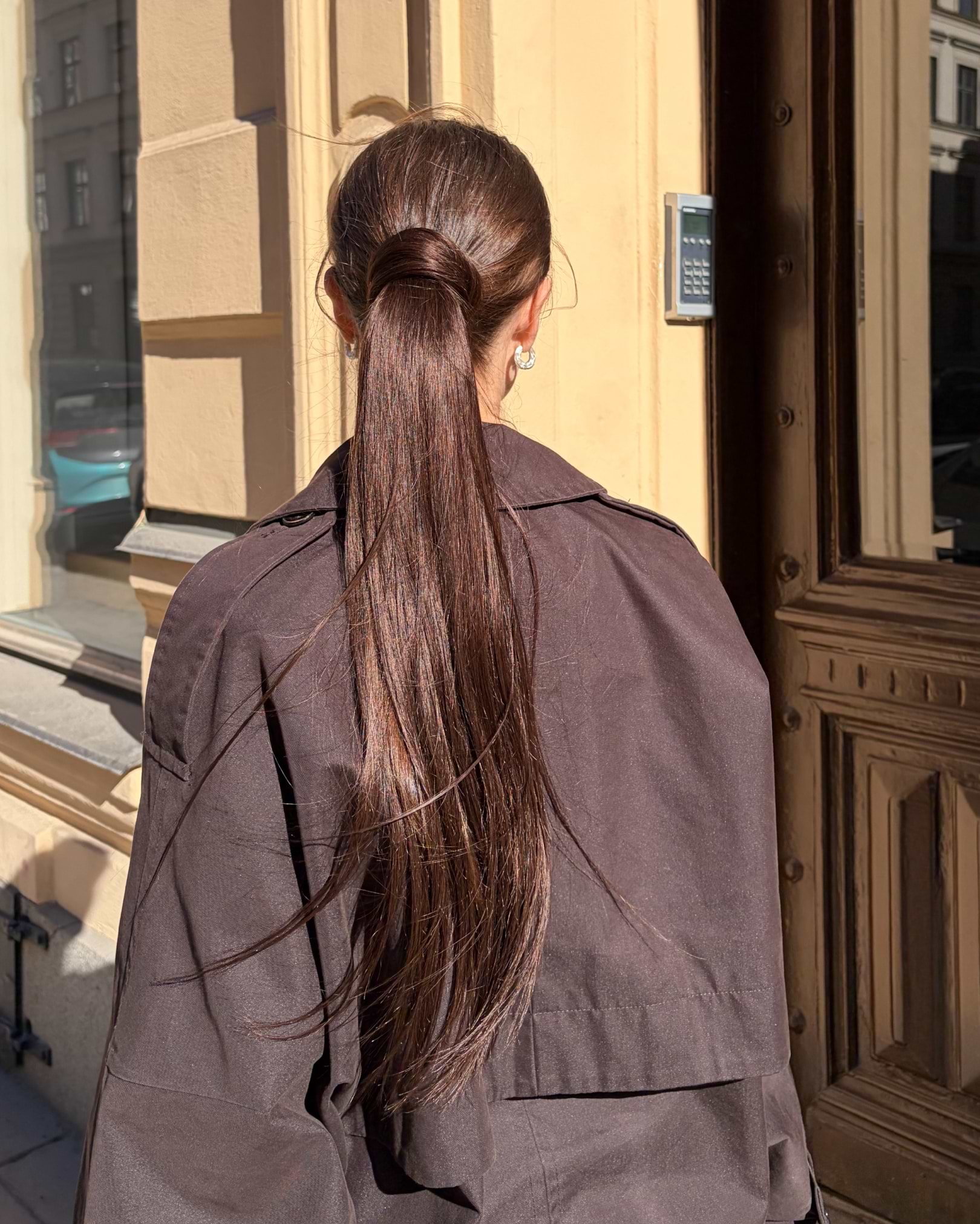 Person with long, straight brown hair in a sleek ponytail, wearing a dark coat, stands outside near a wooden door and beige wall.
