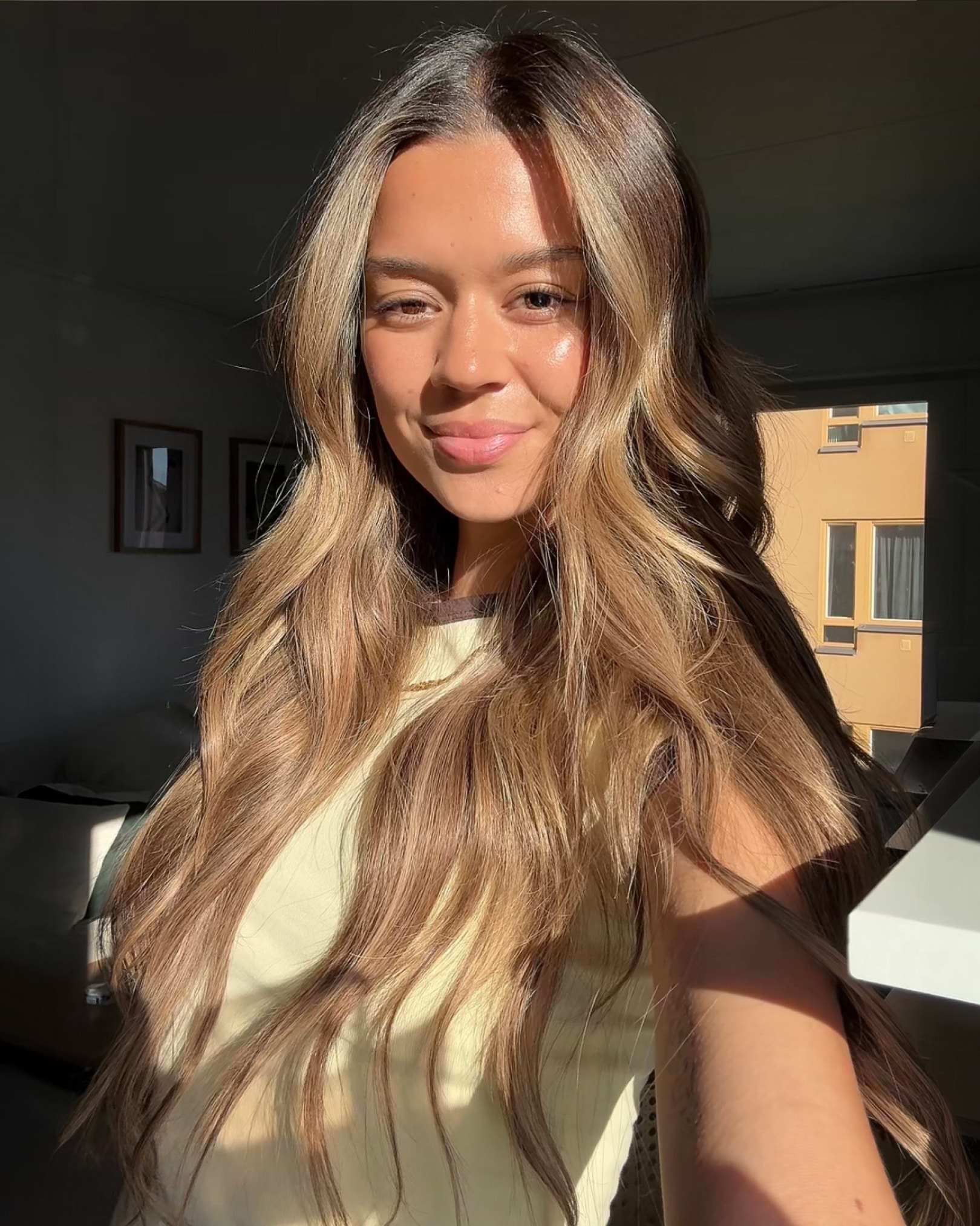 A woman with long, wavy brown hair and a light top, smiling in natural sunlight indoors.