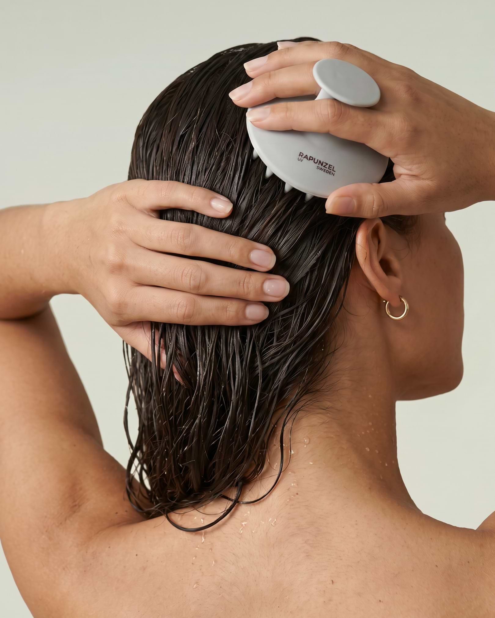 Person using a scalp massager to comb through wet hair, with water droplets visible, viewed from behind.