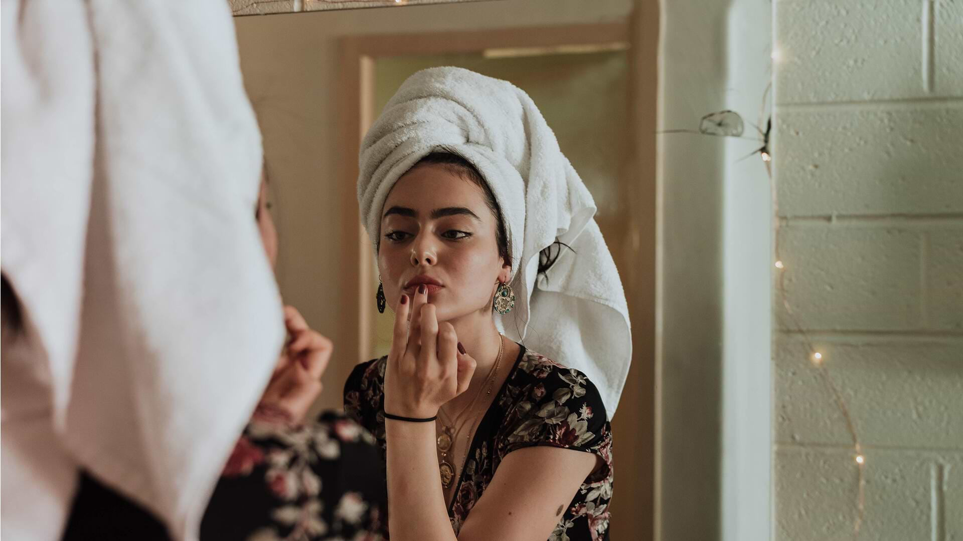 Woman with a towel on her head applies lipstick in front of a mirror, wearing a floral top, in a softly lit room.