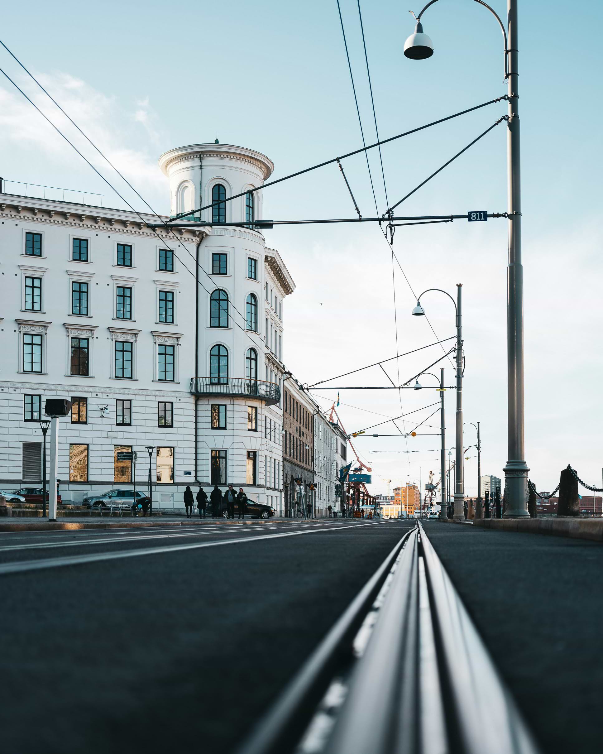 Street view of a city with tram tracks, a historic white building on the left, and streetlights lining the road under a clear blue sky.