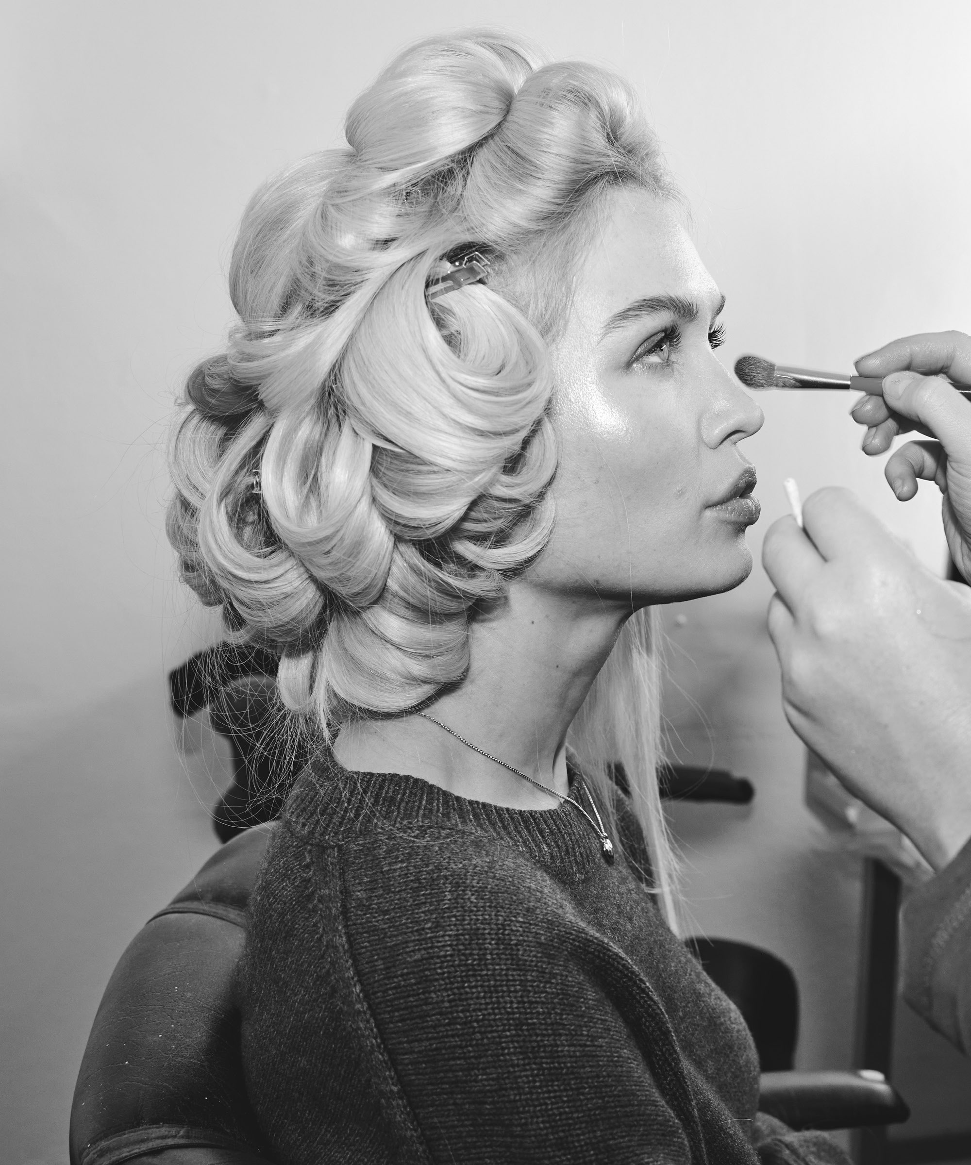 Person with styled hair extensions in rollers, sitting in a chair, having makeup applied with a brush, wearing a sweater.