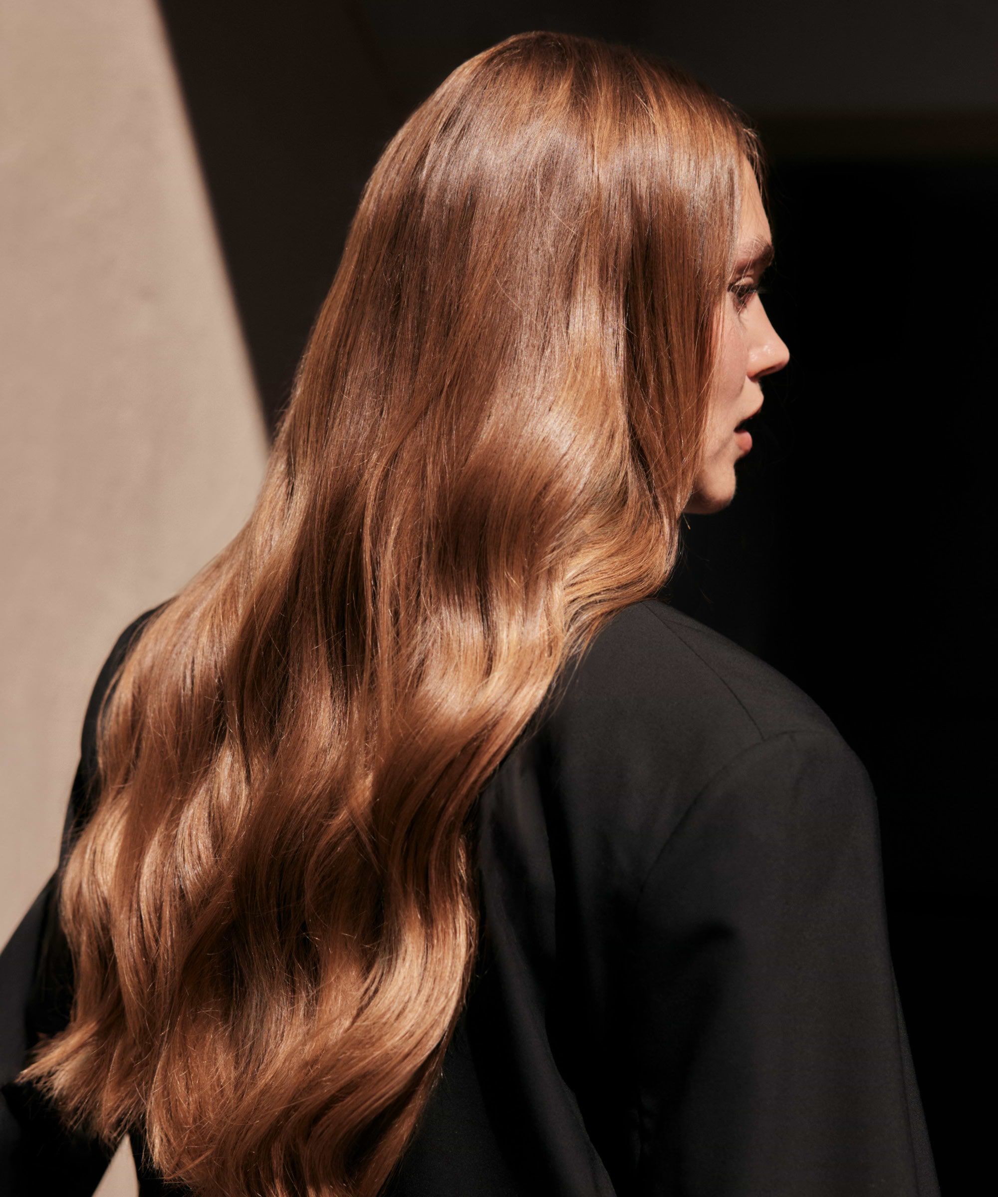 Person with long, wavy, light brown hair extensions, wearing a black jacket, facing away from the camera in natural light.