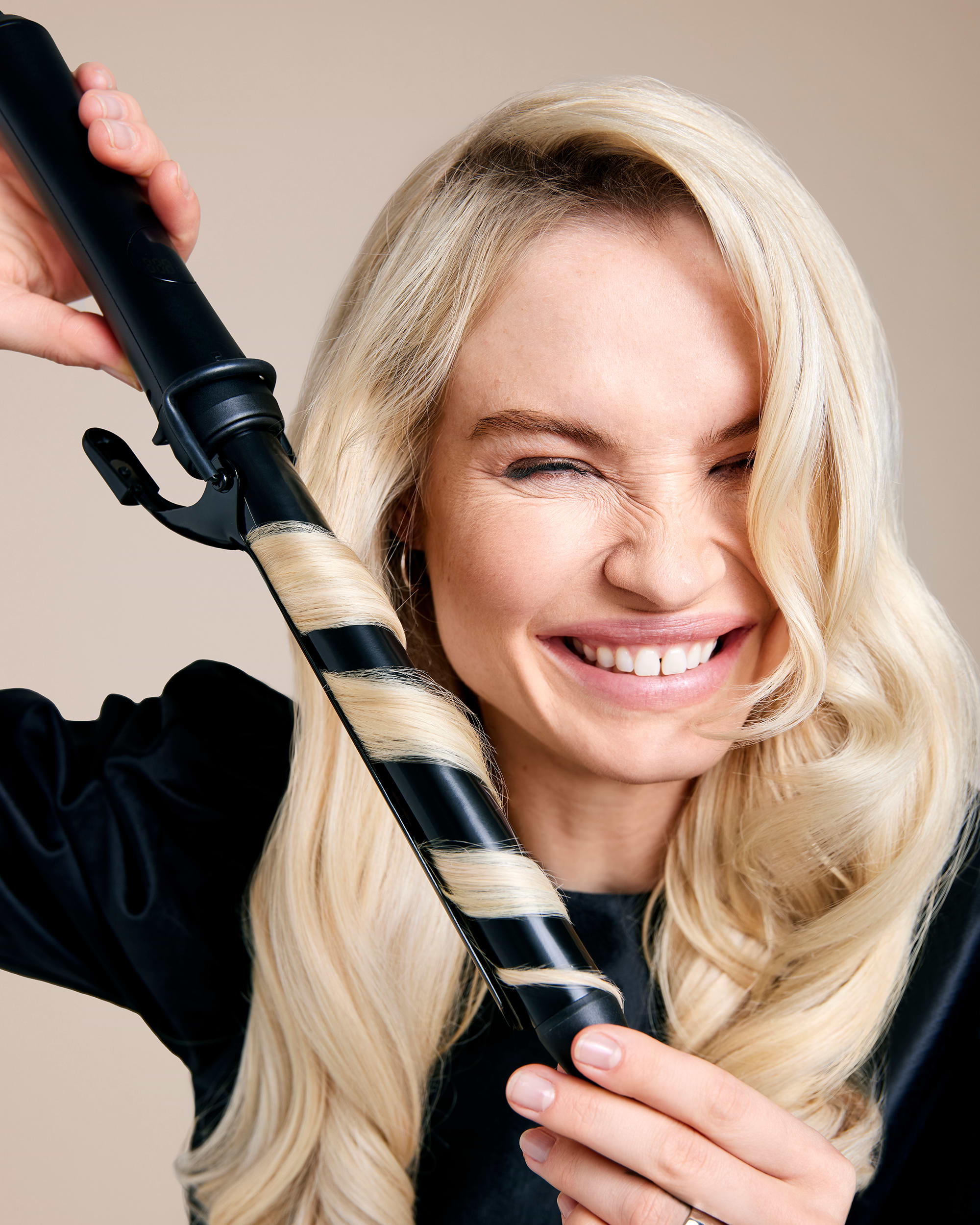 Woman with blonde hair smiling, holding a curling iron with a strand of hair wrapped around it, against a neutral background.
