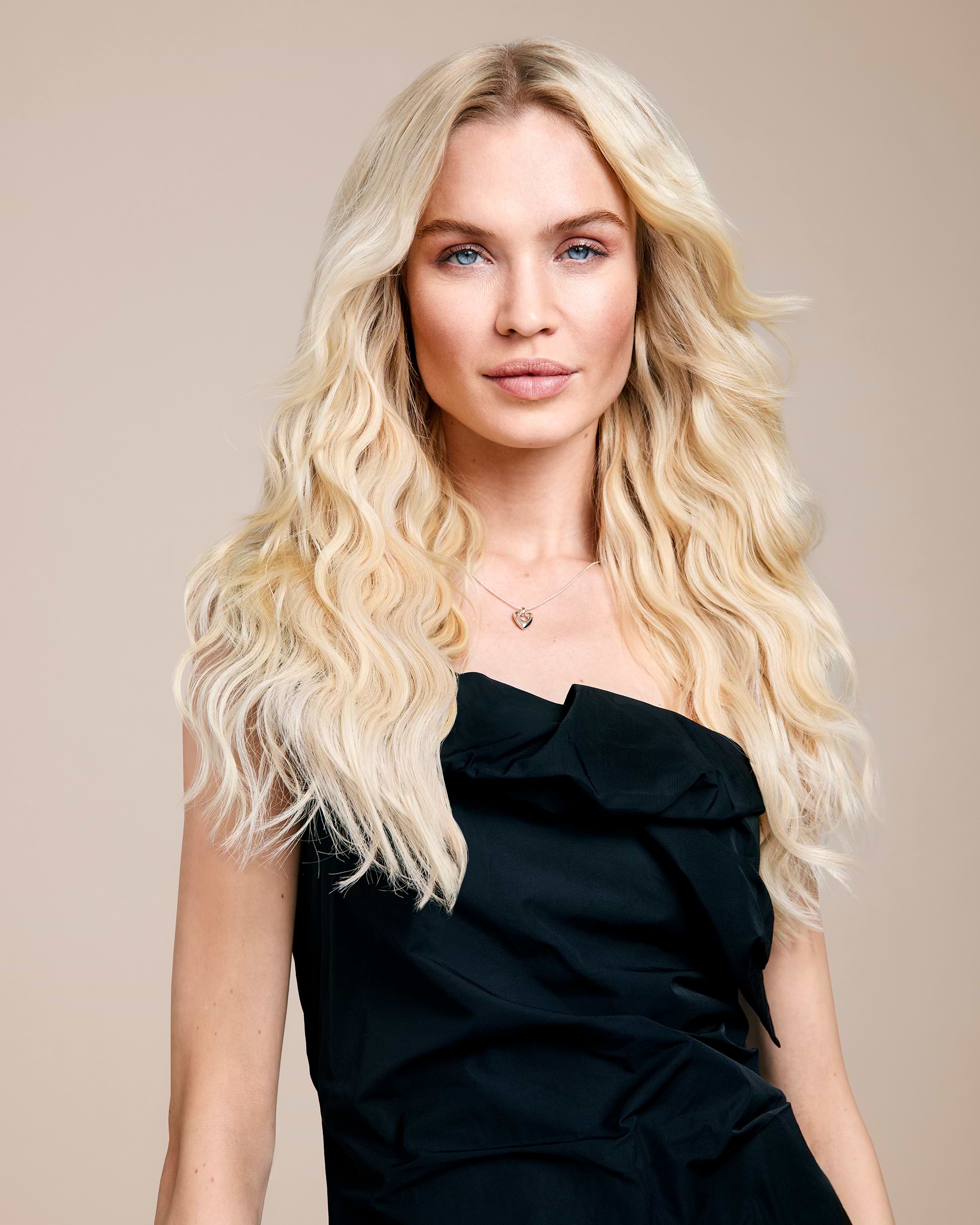 Woman with long wavy blonde hair wearing a black off-shoulder dress, standing against a neutral background.