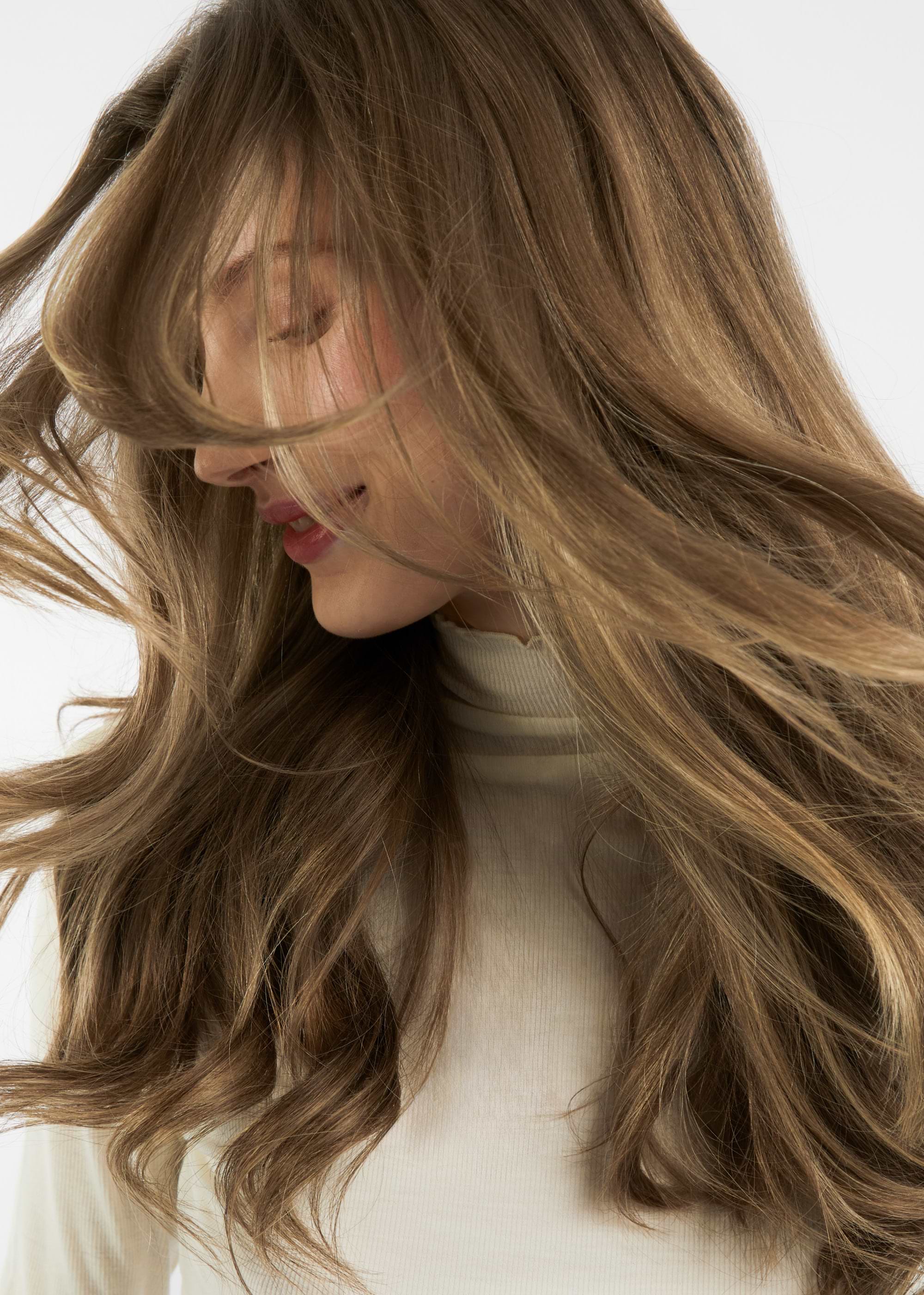 Woman with long, flowing light brown hair extensions, wearing a white turtleneck, smiling and tilting her head to the side against a plain background.