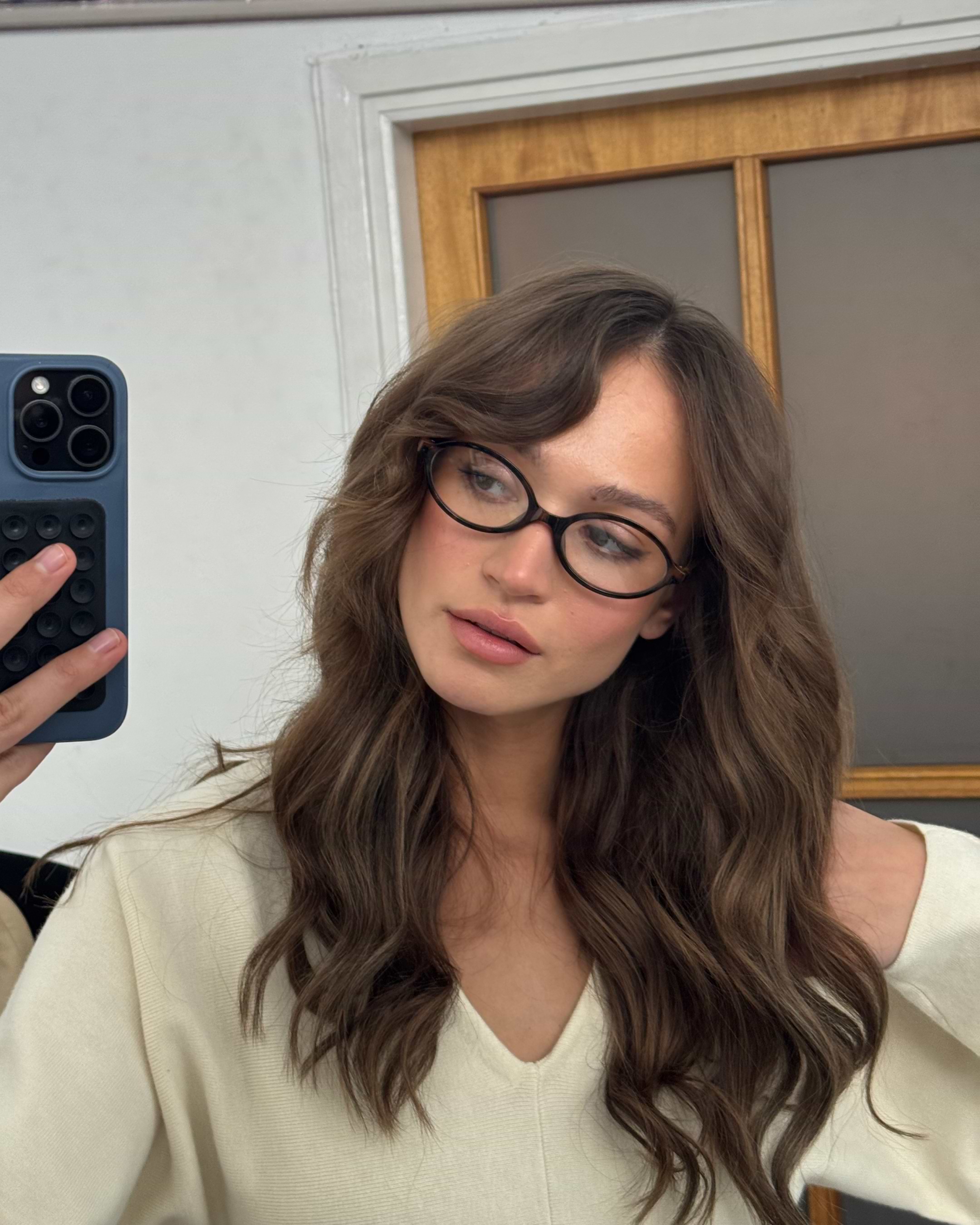 A person with wavy brown hair and glasses takes a mirror selfie in a beige top, holding a smartphone with a black case.