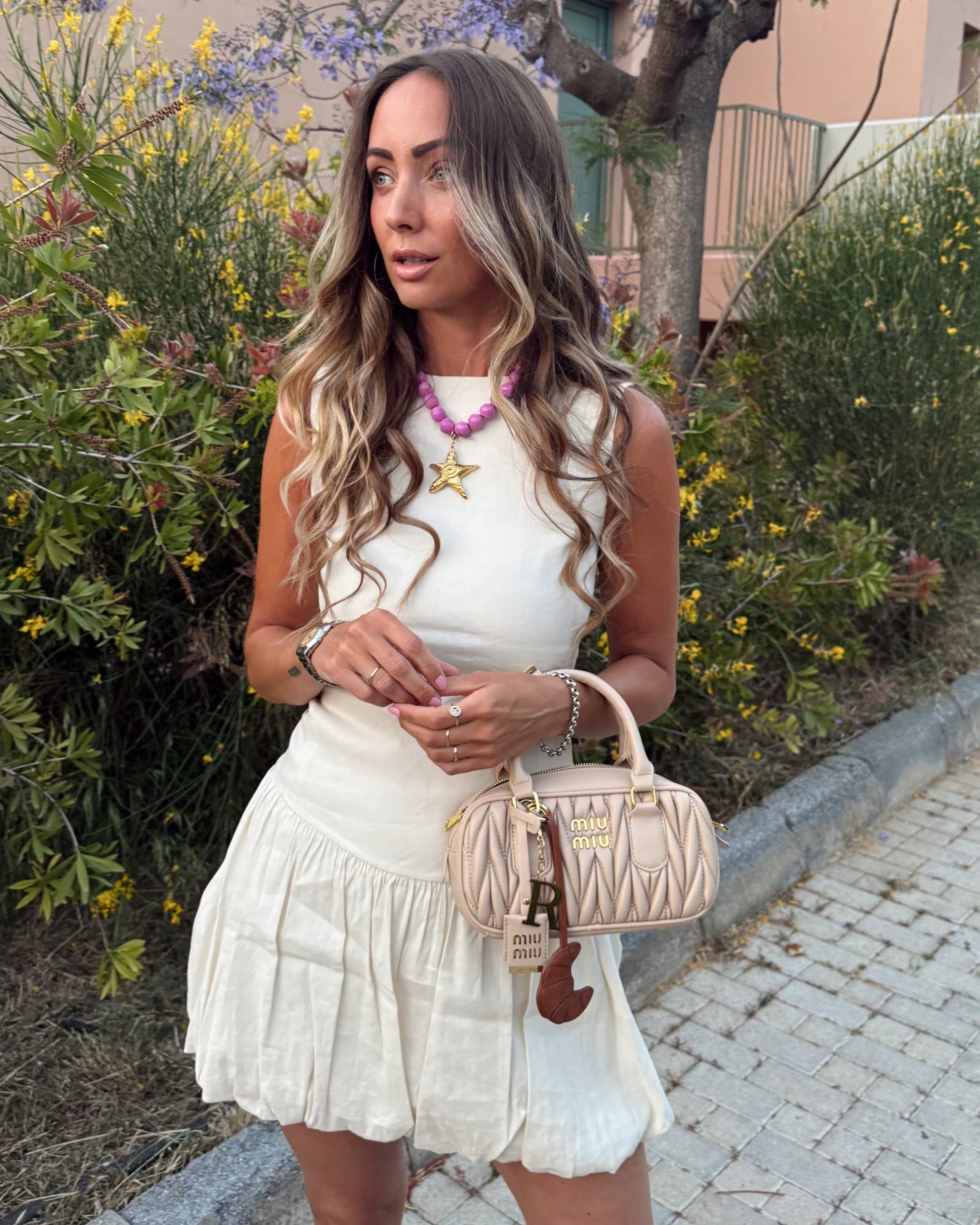 Woman in white dress with star necklace and handbag stands outdoors on a paved path, surrounded by greenery and yellow flowers.