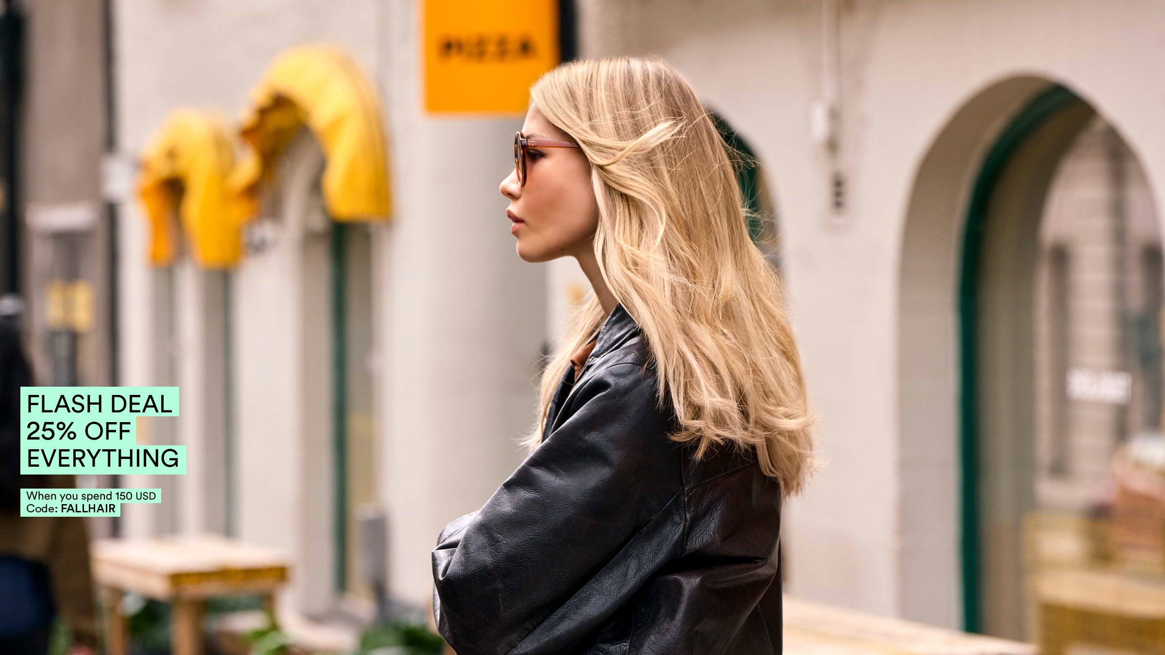 A woman with long blonde hair wearing sunglasses and a black jacket stands outside near a store. A flash deal sign is visible.