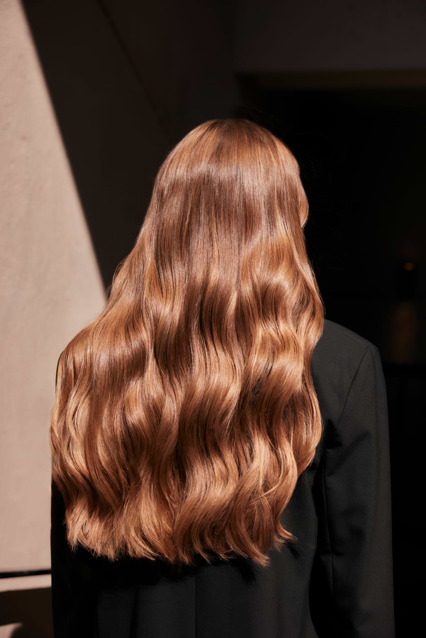 A person with long, wavy, light brown hair extensions stands with their back to the camera, wearing a black top.