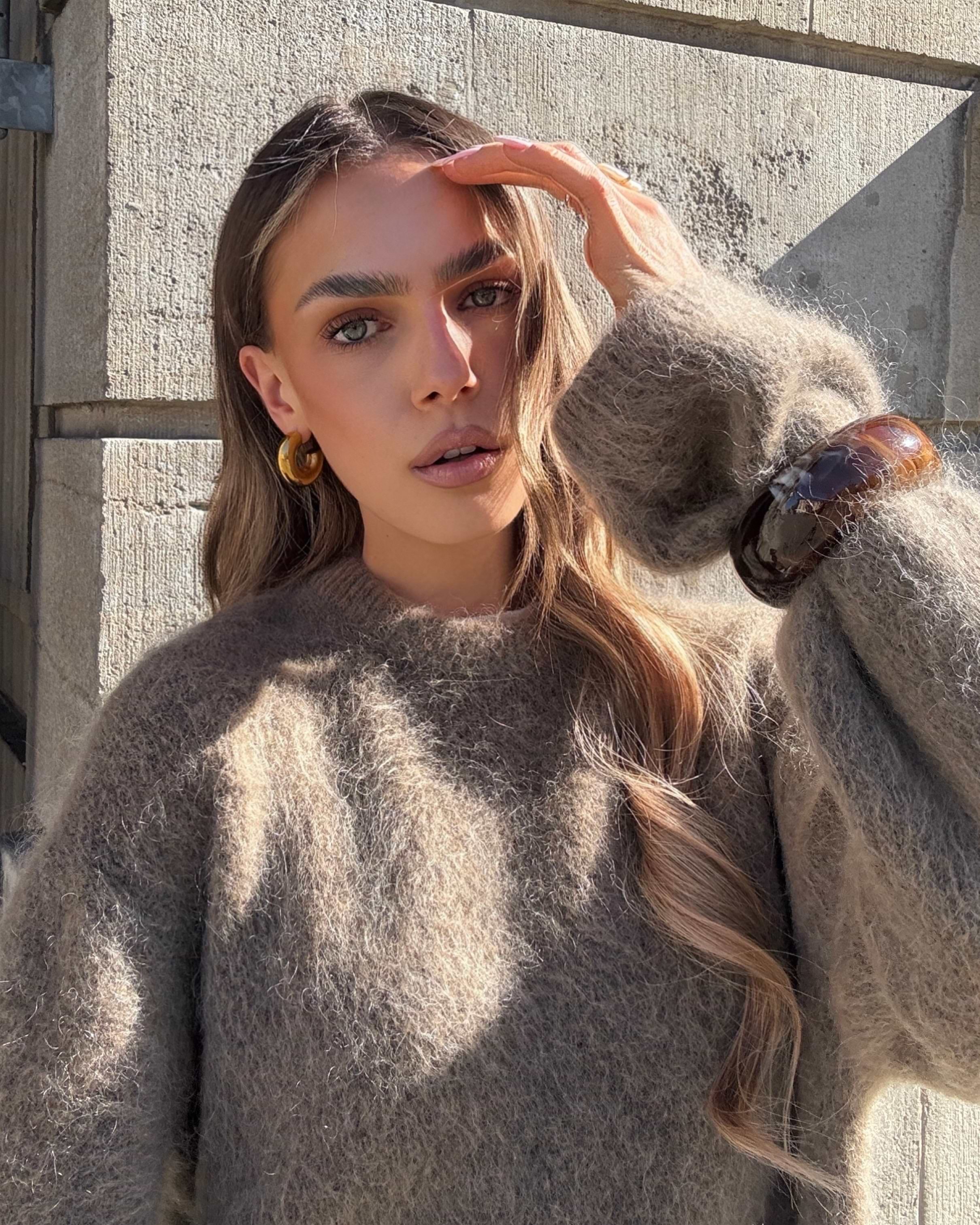 Woman with long hair wearing a cozy brown sweater and large bracelet, standing outdoors with a hand shielding the sun from her face.