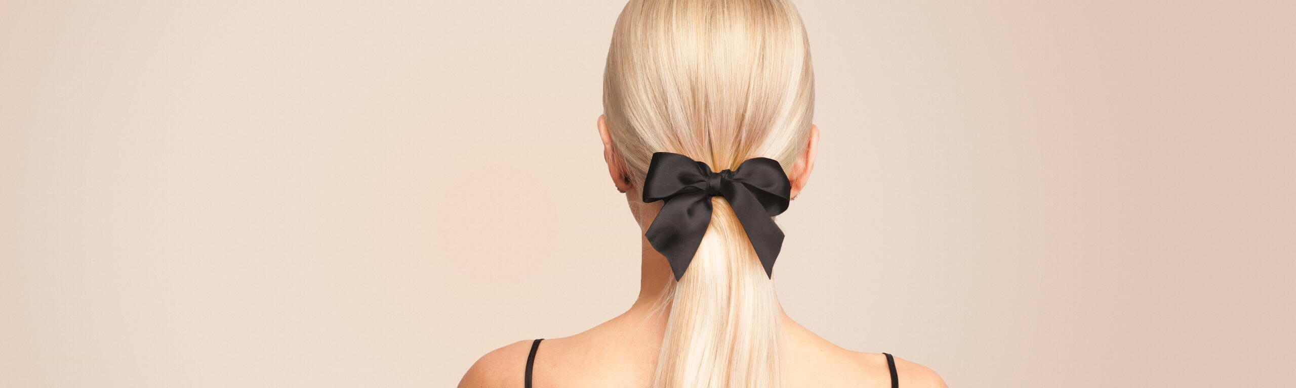 Rear view of a person with long blonde hair extensions tied in a ponytail, adorned with a large black bow, against a neutral background.