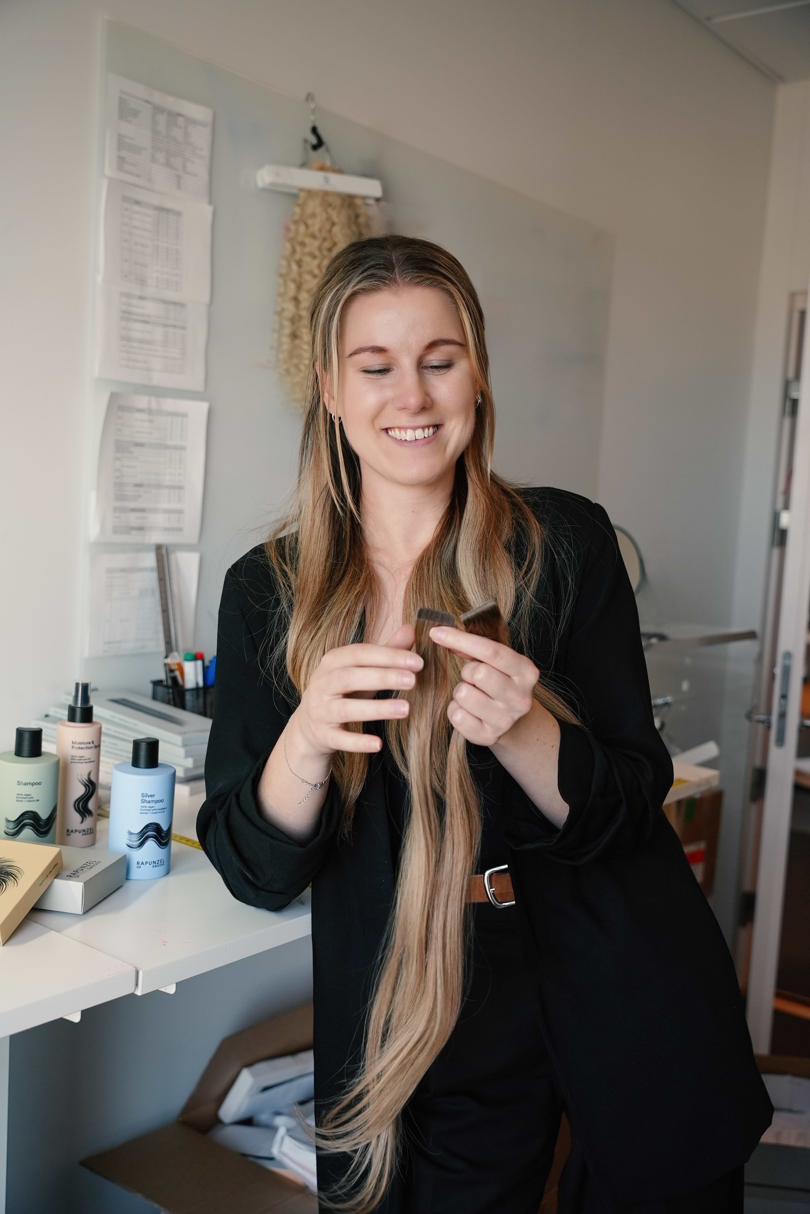 Woman with long hair extensions in a black outfit smiles while holding a hair extension in an office with hair products on a desk.