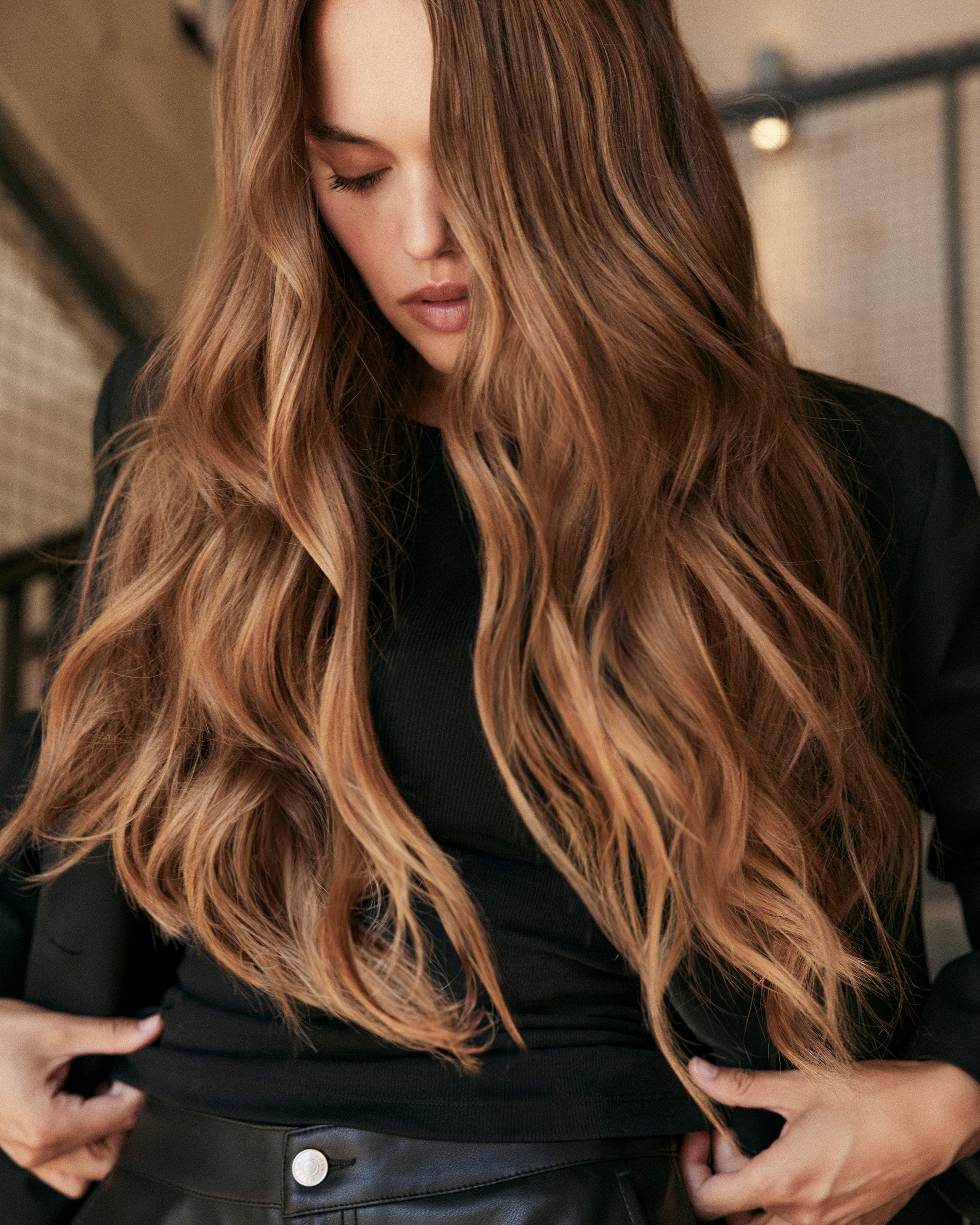 Woman with long, wavy brown hair extensions wearing a black top and jacket, looking down, standing indoors with a neutral background.