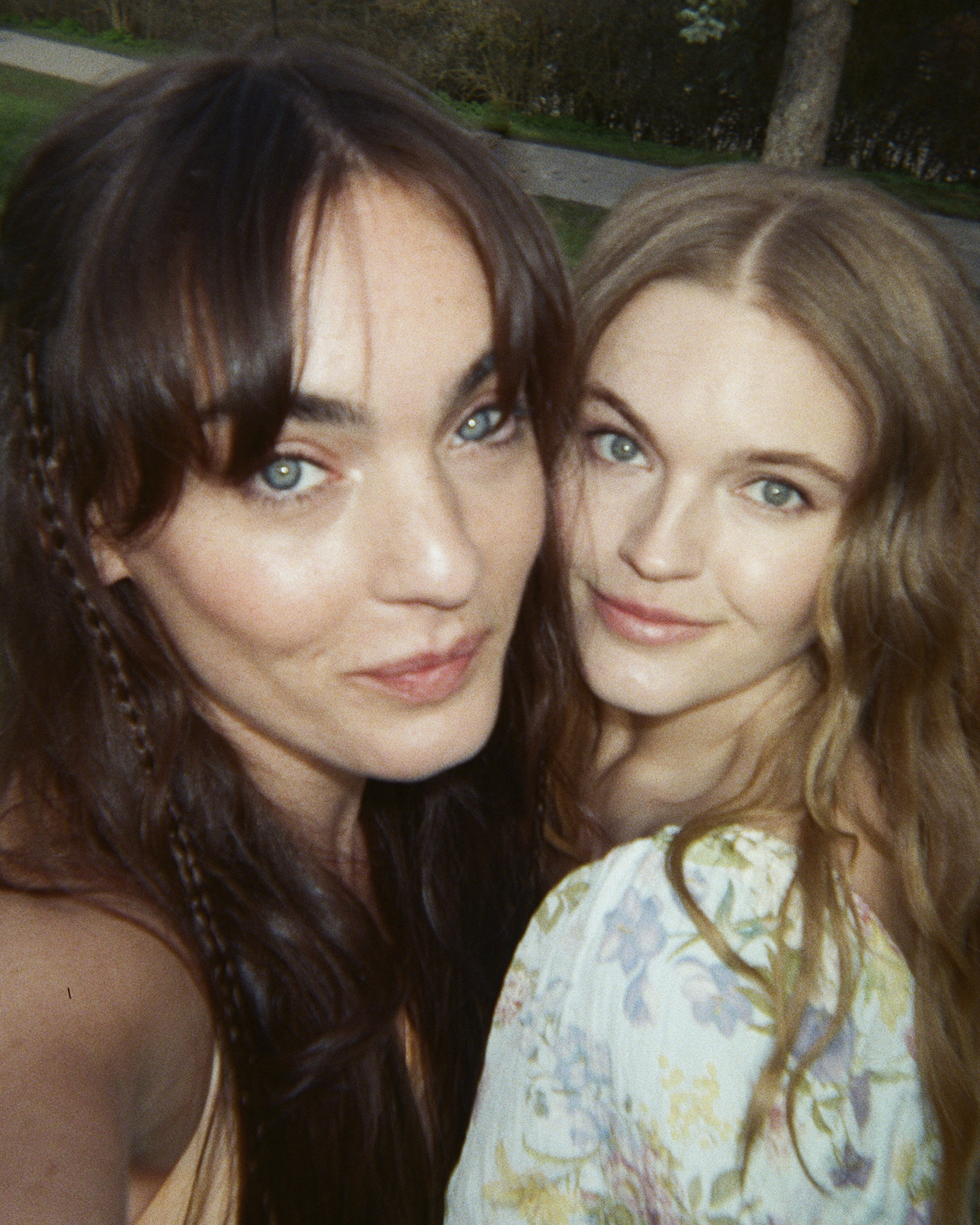 Two women smiling for a selfie outdoors, one with long dark hair and the other with wavy blonde hair extensions, both wearing light-colored tops.