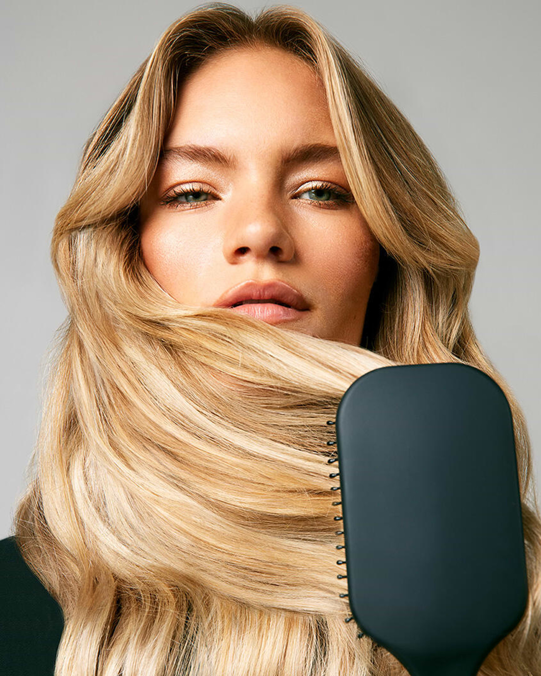 Woman with long blonde hair holding a black paddle brush in front, hair cascading over the brush. Neutral background.