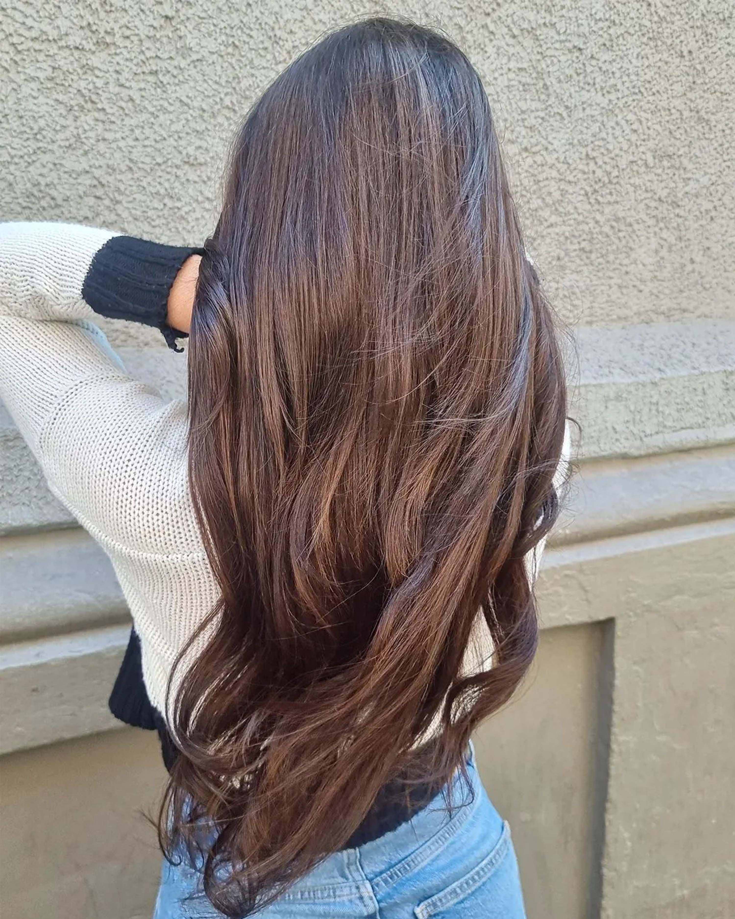 Person with long, wavy brown hair wearing a cream and black sweater and jeans, standing against a textured wall with their back to the camera.