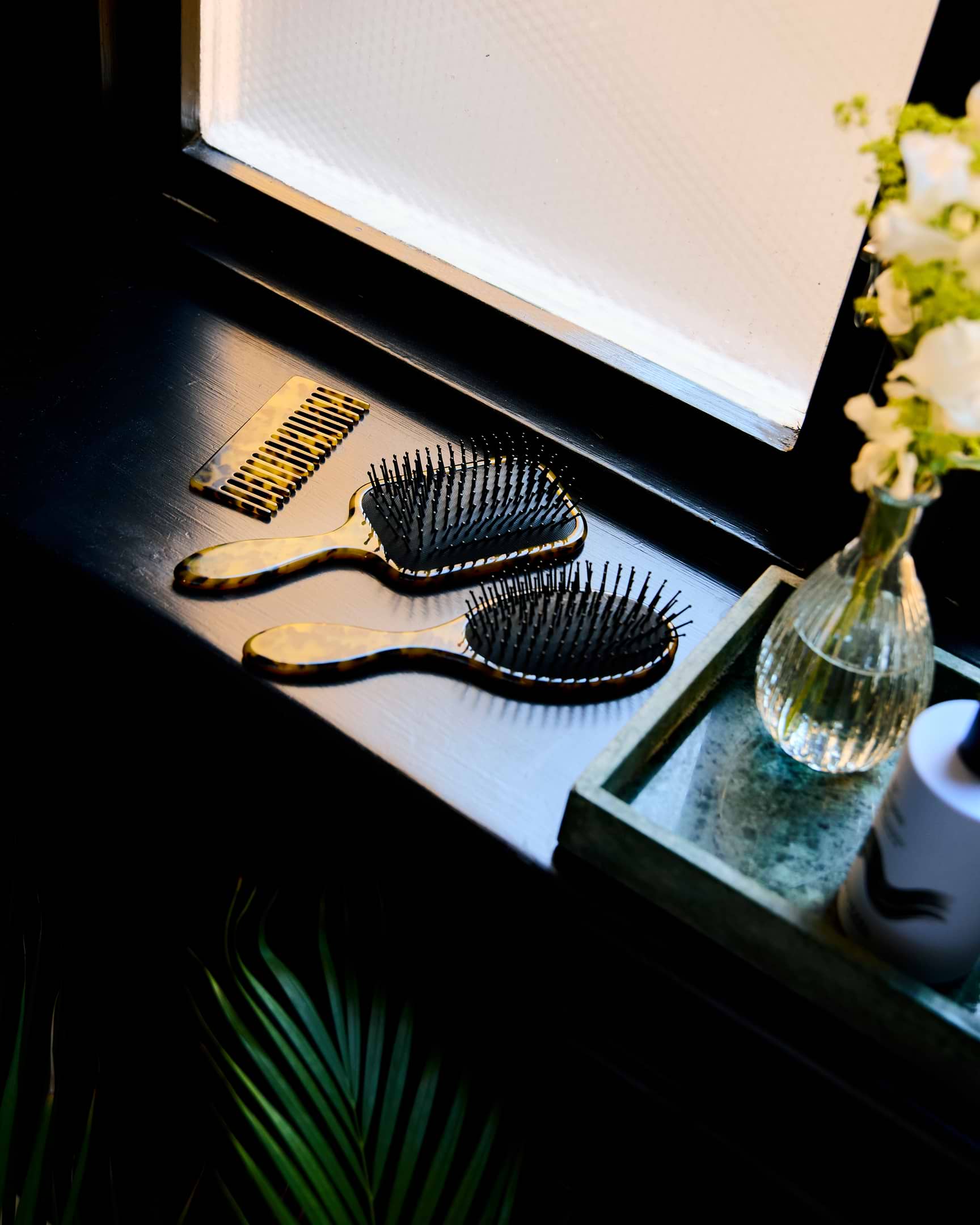 Two hairbrushes with tortoiseshell handles on a windowsill, next to a small vase with white flowers and a green tray.
