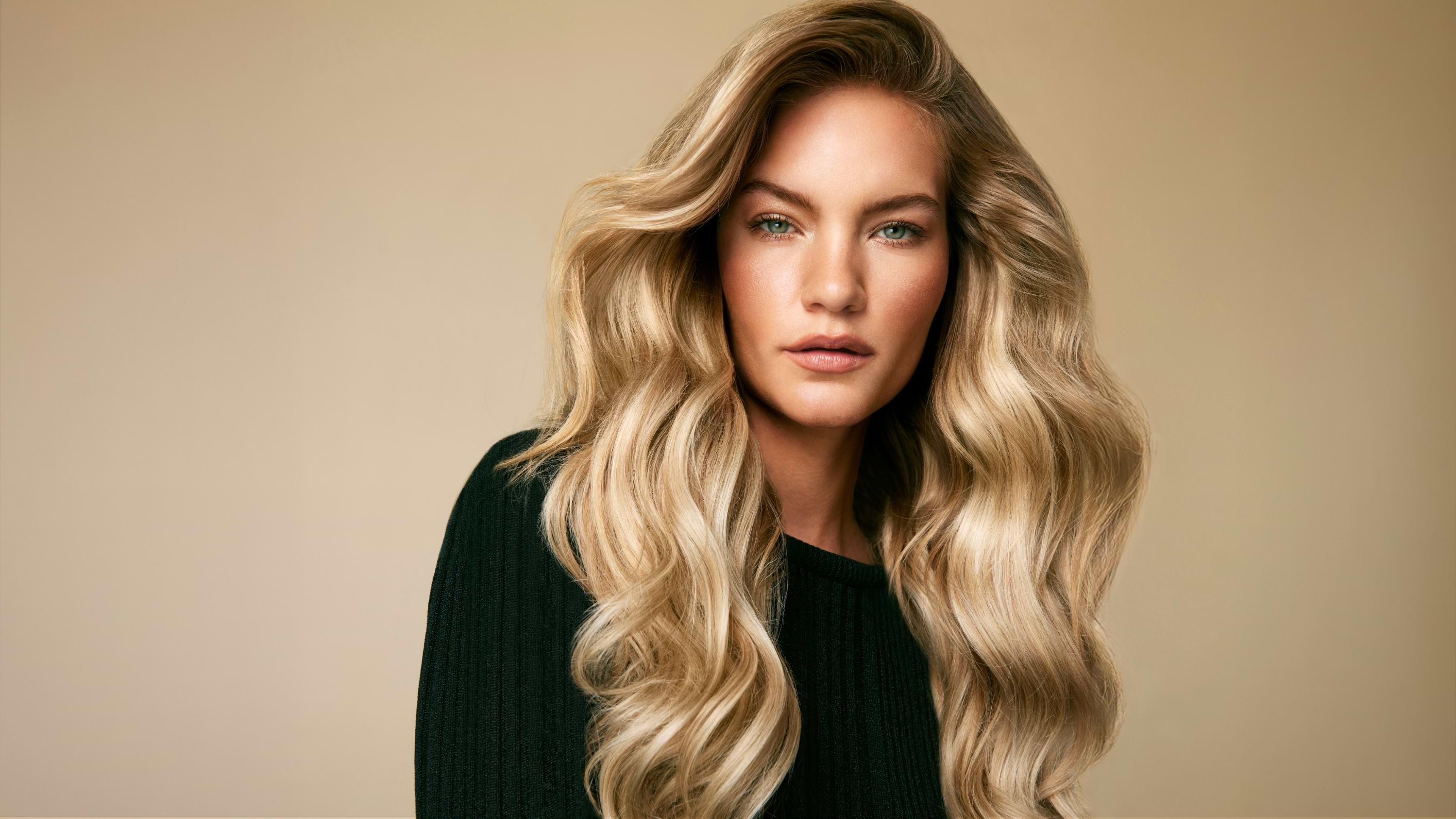 A person with long, wavy blonde hair wearing a black sweater, looking directly at the camera with a neutral expression against a beige background.