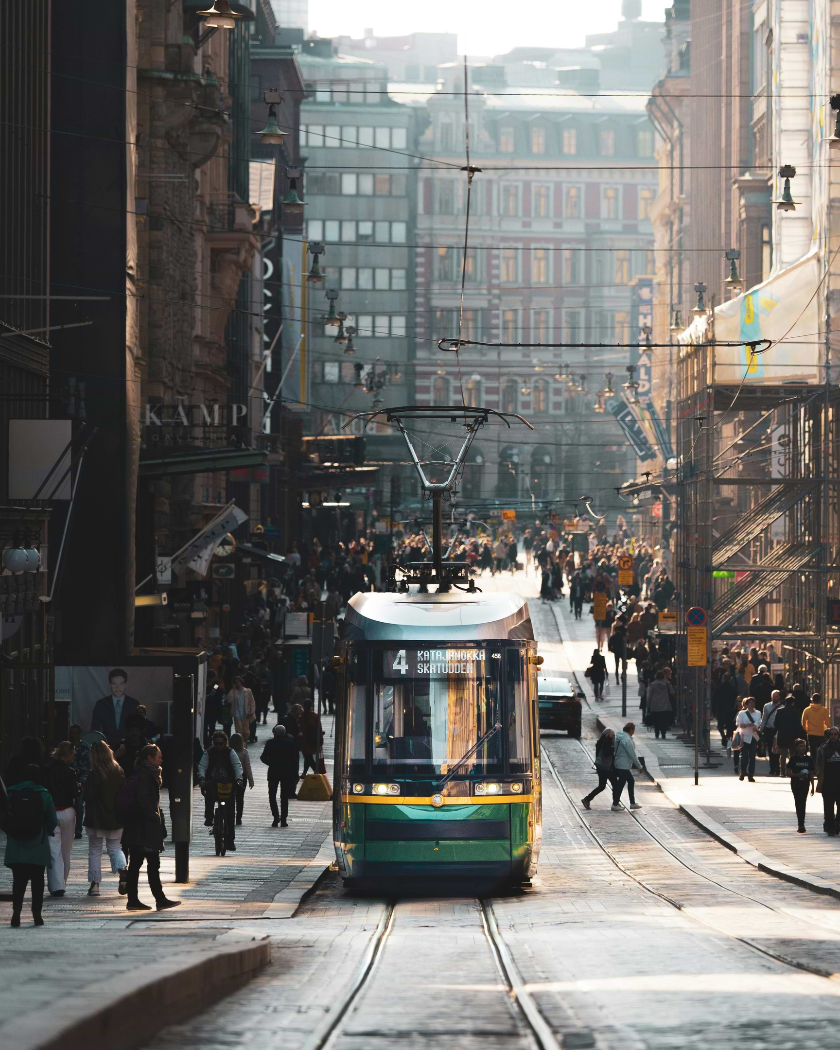 A green tram travels down a bustling city street lined with historic buildings and crowded with pedestrians.