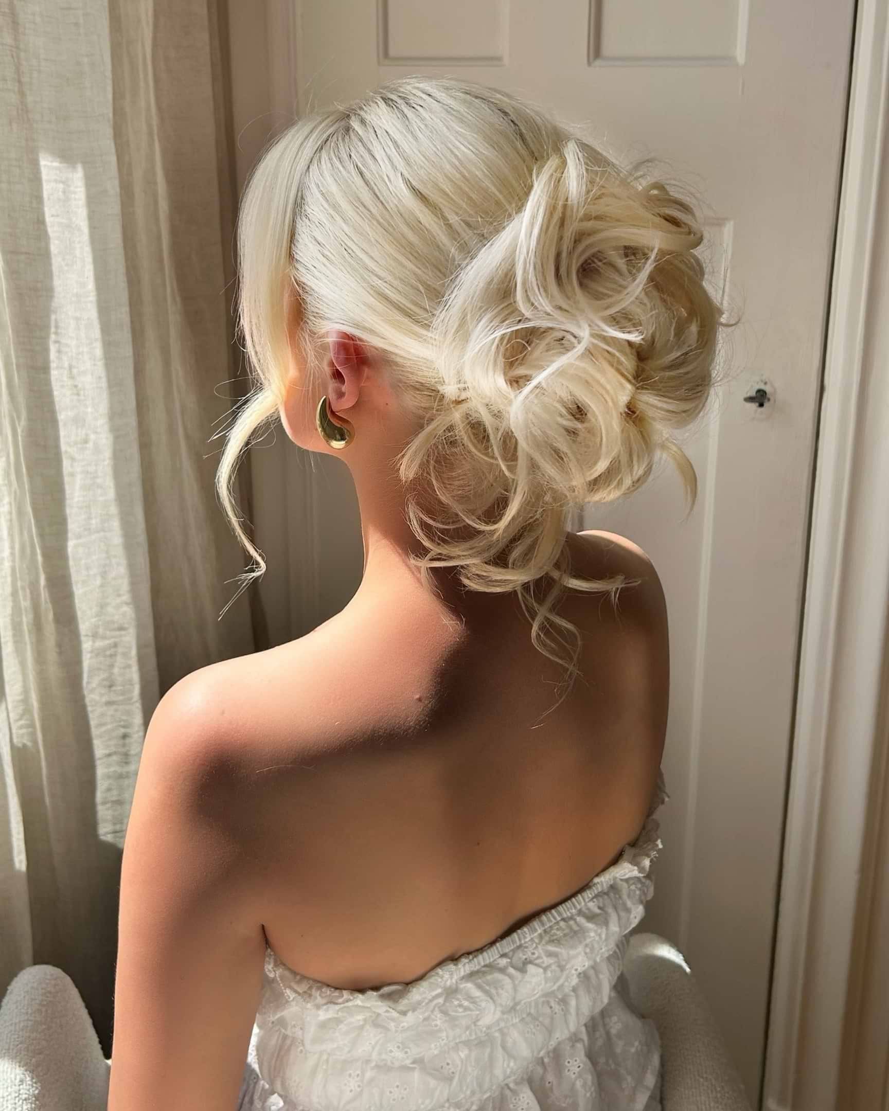Woman with blonde hair in an elegant updo, wearing a strapless white dress, sitting in soft light by a window.