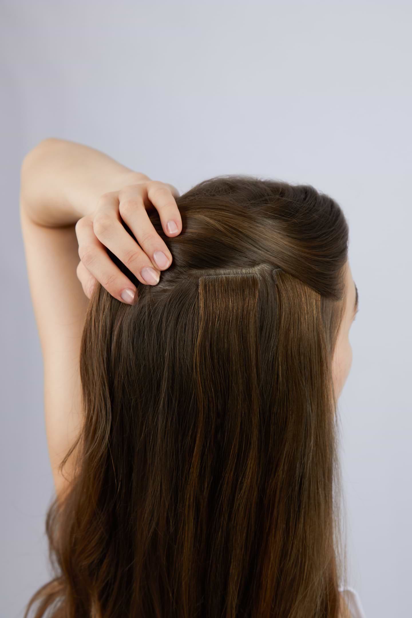 Person with long brown hair, showing a hidden hair extension by lifting the top layer of hair with their hand, against a plain background.