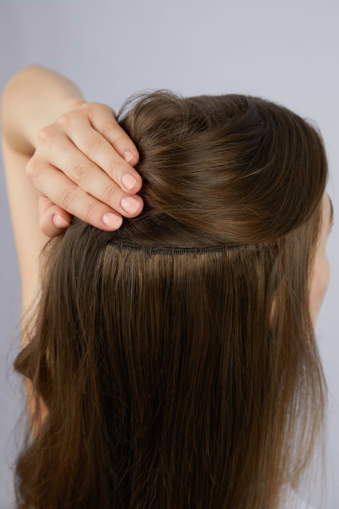 A person demonstrates a hair extension application by lifting brown hair to reveal the attachment near the scalp.